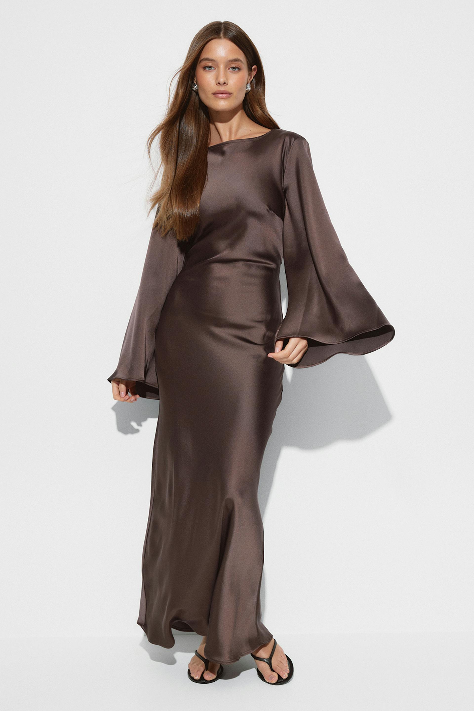 Robe longue en satin brun chocolat avec encolure bateau, taille ajustée et manches cloche volumineuses, à porter avec des sandales thong noires.