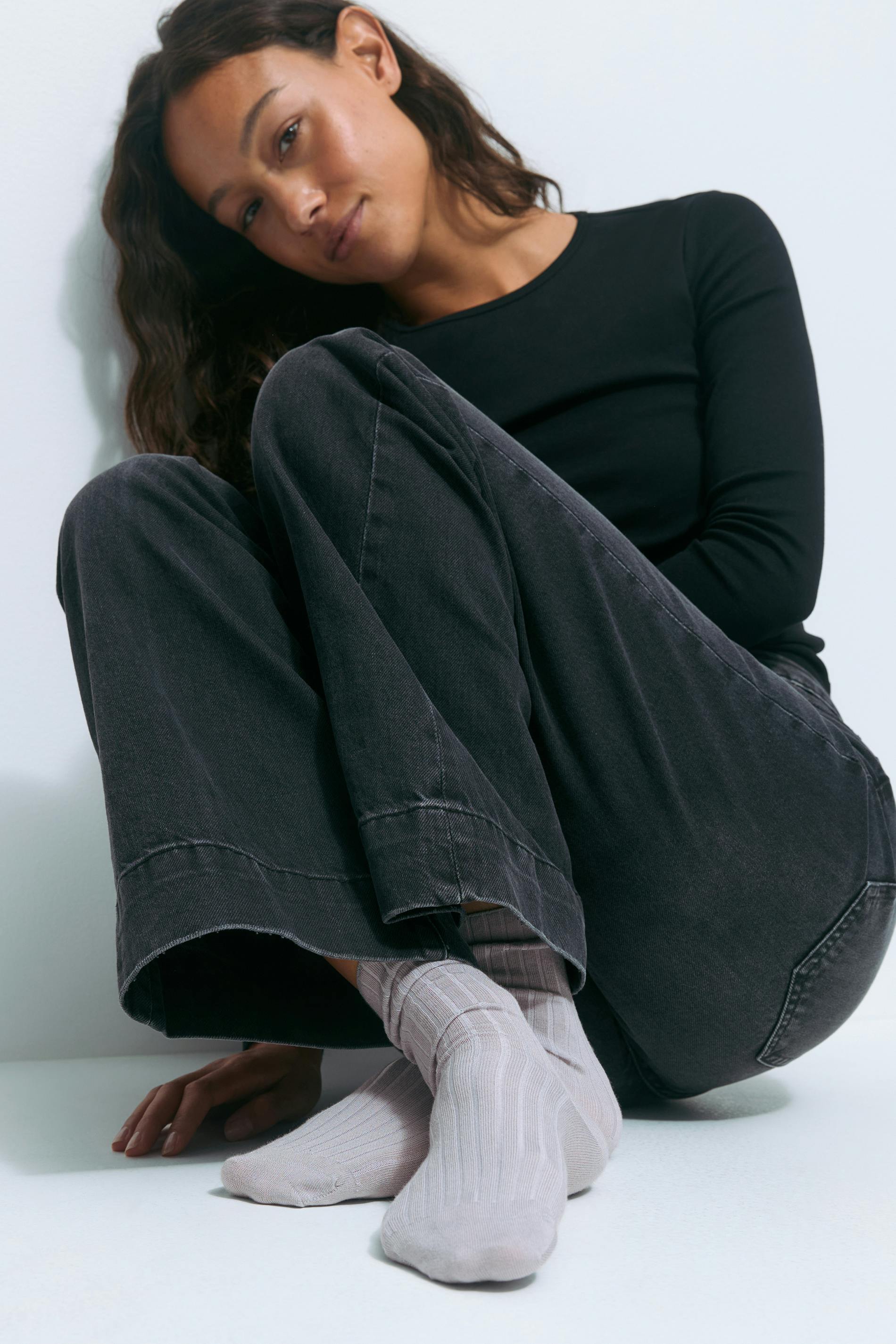 Frau trägt ein schwarzes langärmeliges Oberteil mit Rundhalsausschnitt, verblasste anthrazitfarbene Jeans mit weitem Bein und hellgraue gerippte Socken mit Rundhalsausschnitt.