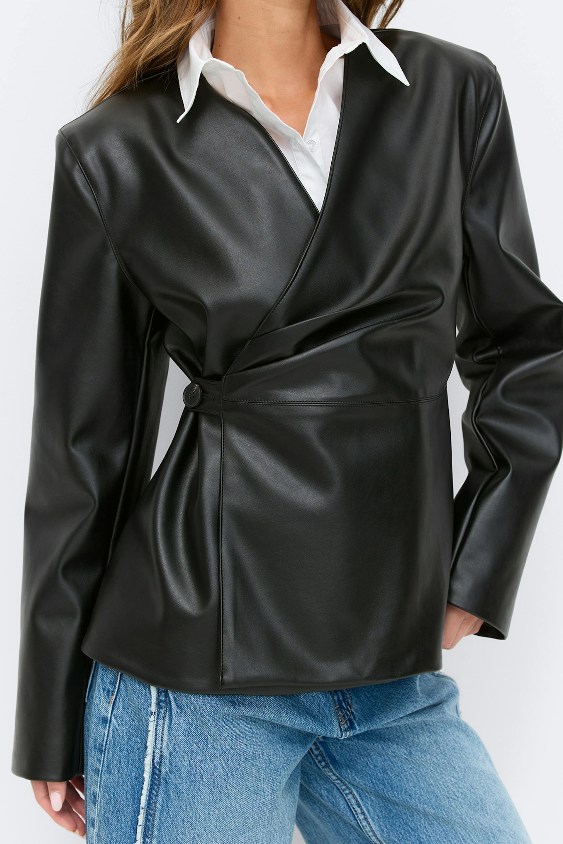 Musta keinonahkainen kietaisutakki, jossa on yhden napin vyötärösuljenta, valkoisen kauluspaidan ja vaalean sinisten farkkujen päällä.