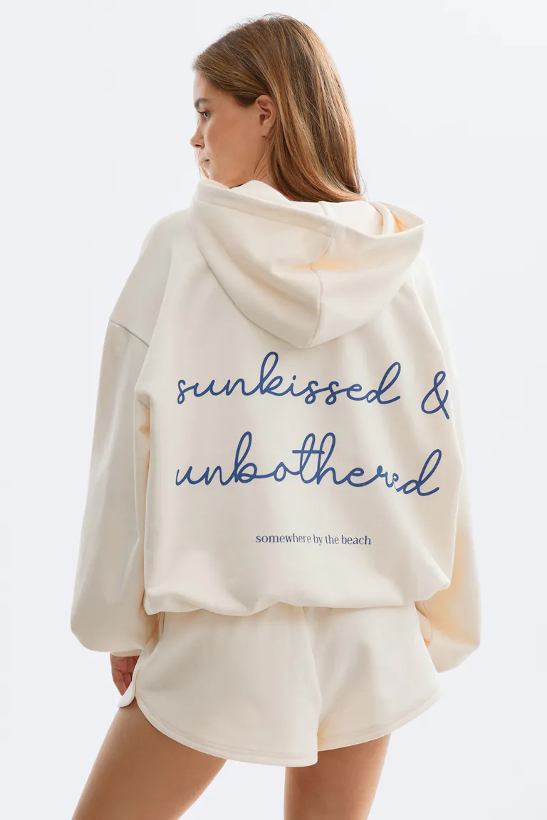 Cremefarbenes, übergroßes Kapuzensweatshirt mit blauem Schriftzug auf der Rückseite mit der Aufschrift "sunkissed & unbothered", gepaart mit passenden cremefarbenen, locker sitzenden Shorts.