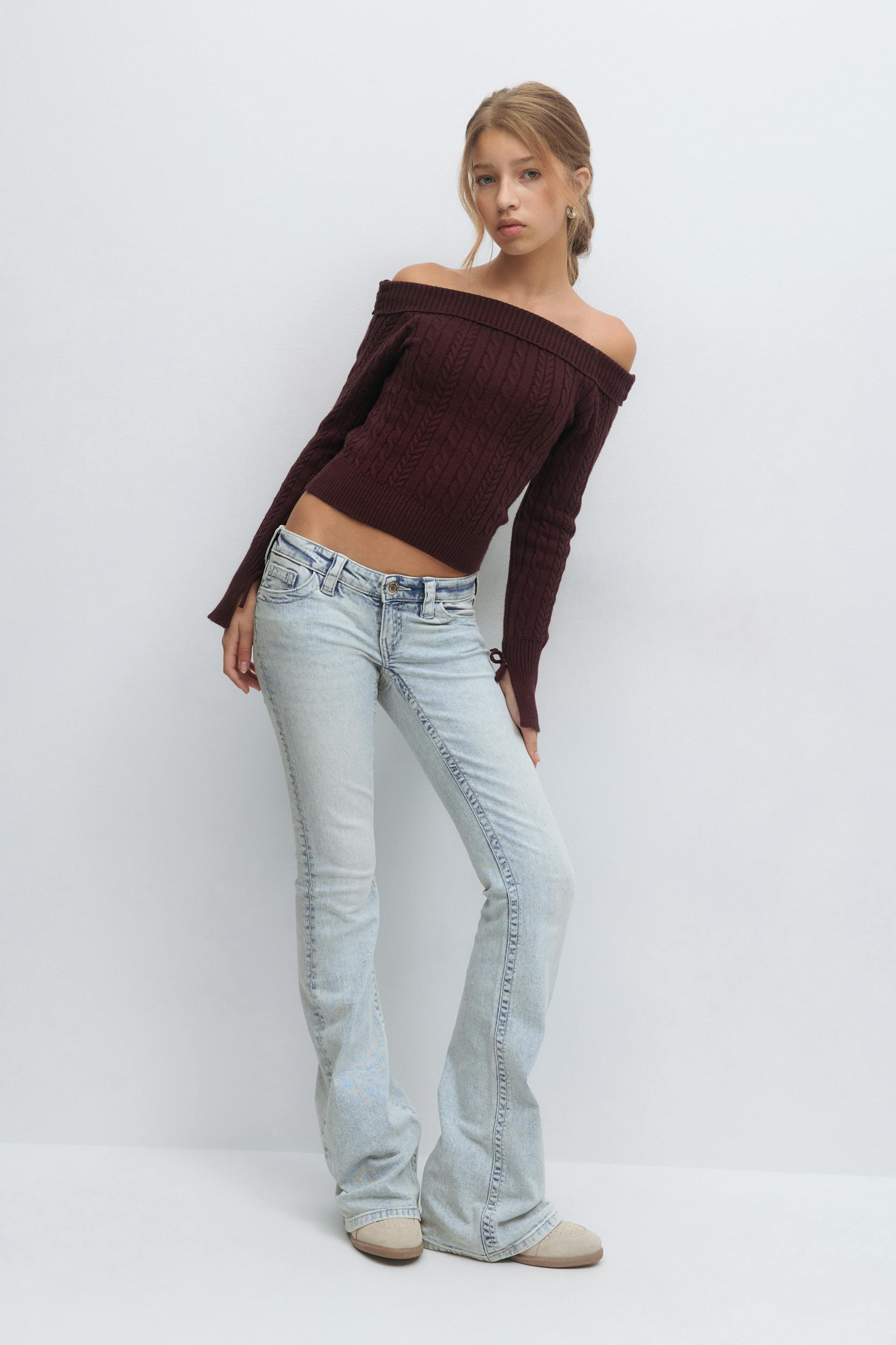 Burgundy off-the-shoulder cable‑knit cropped sweater, light‑wash low‑rise flared jeans, and beige slip‑on shoes.