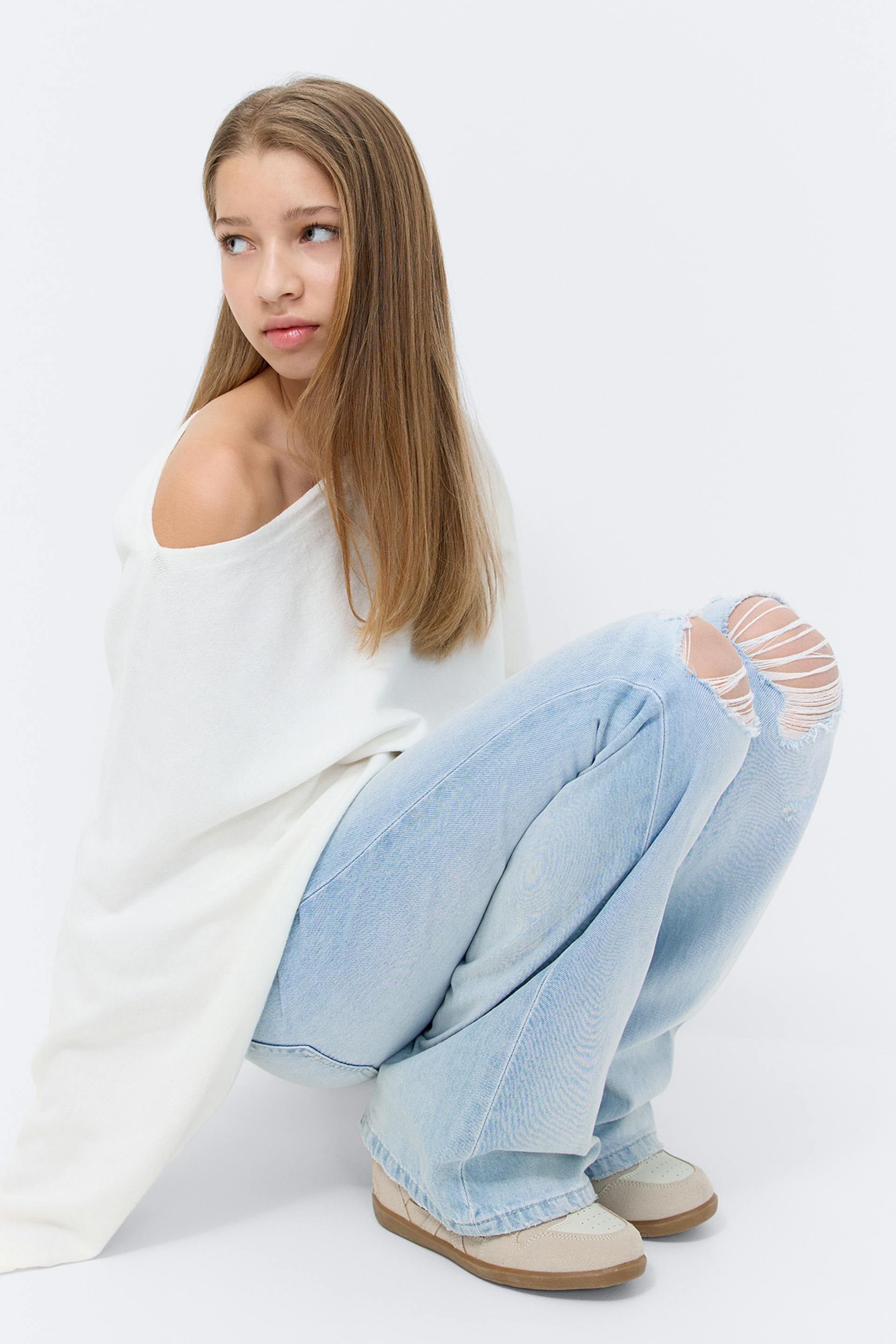 Off-white off-the-shoulder oversized knit sweater, light-wash wide-leg jeans with ripped knees, and beige low-top sneakers with gum soles.