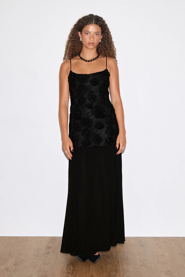 Schwarzes Abendkleid mit dünnen Spaghetti-Trägern, einem floral strukturierten, taillierten Mieder und einem langen, fließenden Rock; kombiniert mit einer schwarzen Perlenkette, kleinen Ohrsteckern und spitzen schwarzen Absätzen.