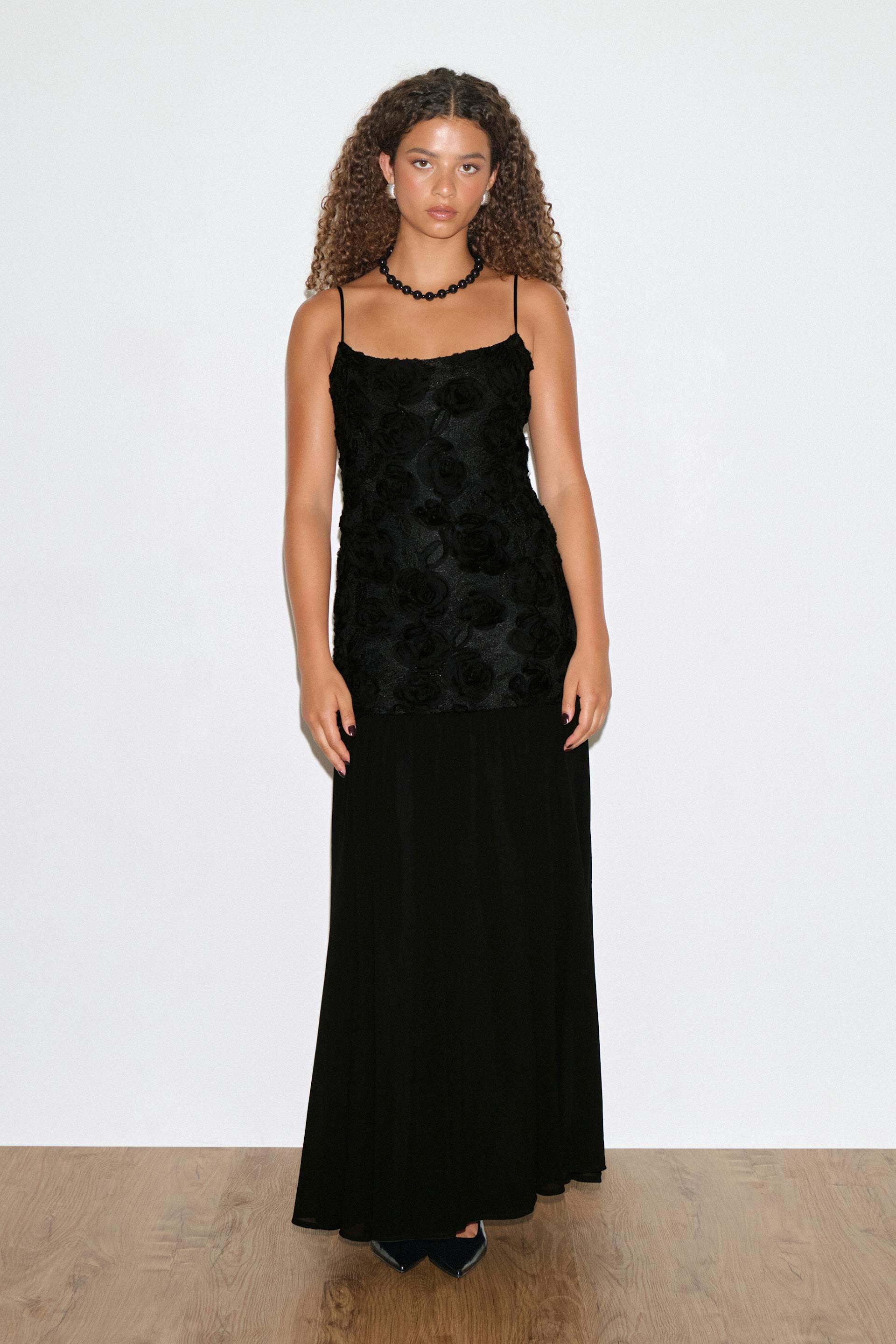 Black evening gown with thin spaghetti straps, a floral-textured fitted bodice and a long flowing skirt; styled with a black beaded necklace, small stud earrings and pointed black heels.