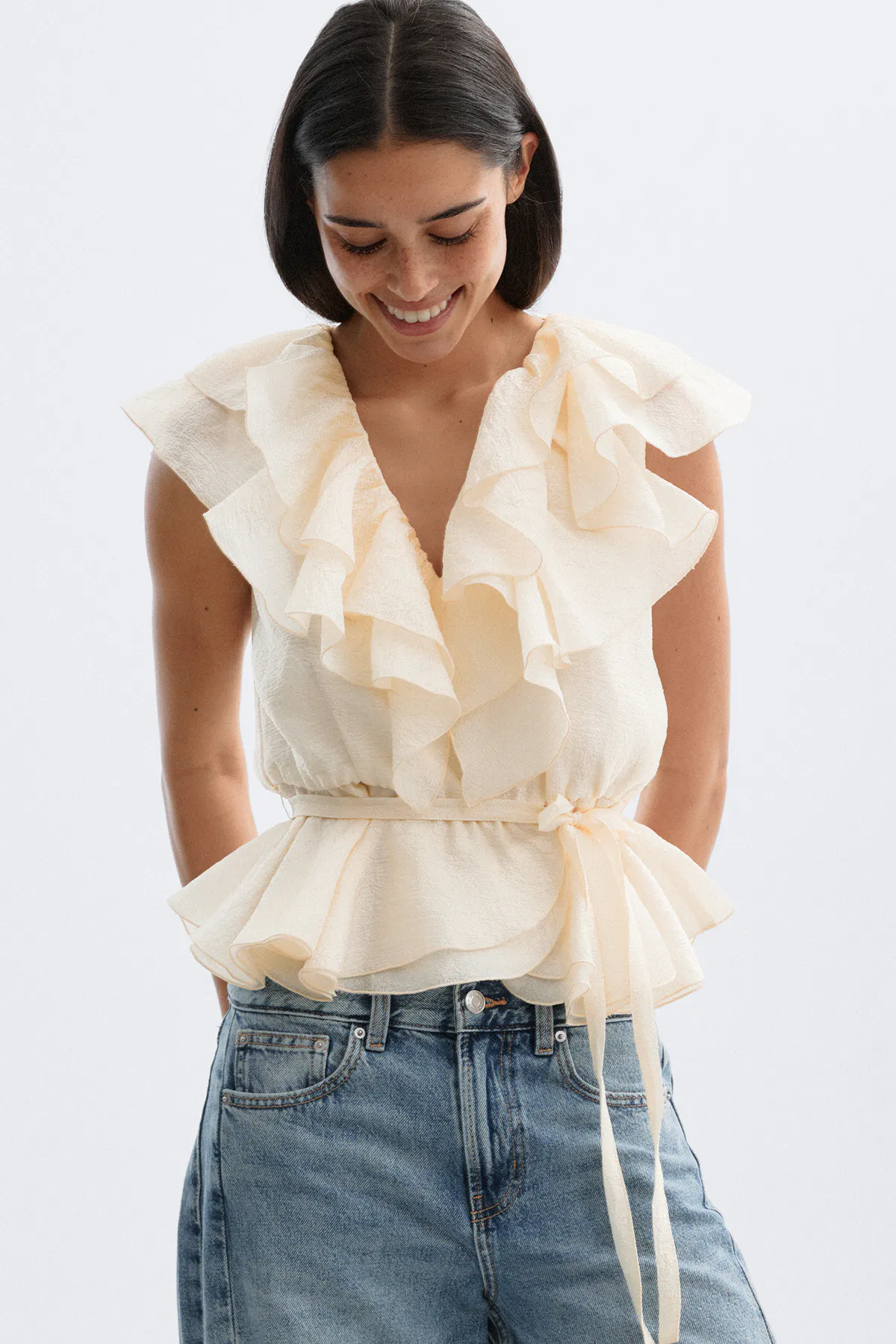 Ærmeløs cremefarvet bluse med flæse i V-udskæring, lagdelt peplum og selvbinderbælte i taljen, sat sammen med lysvaskede højtaljede jeans.