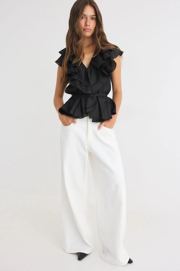 Sleeveless black ruffled blouse with wide-leg white trousers and black pointed shoes.