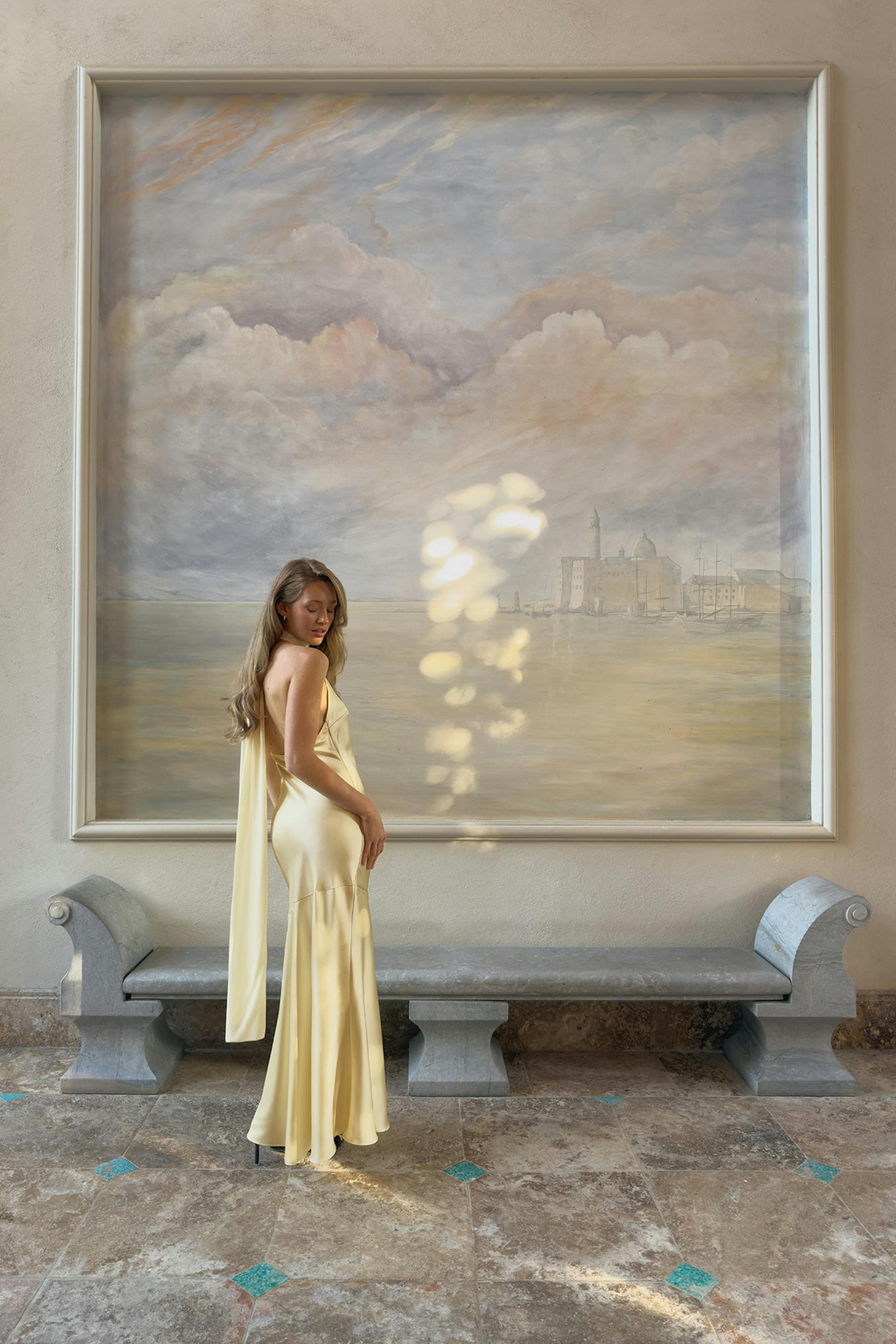 Golvlång aftonklänning i ljusgul satin med enkelt draperat axelskärp, figursydd sjöjungfrusilhuett och utsvängd fåll, stylad med svarta stilettklackar.