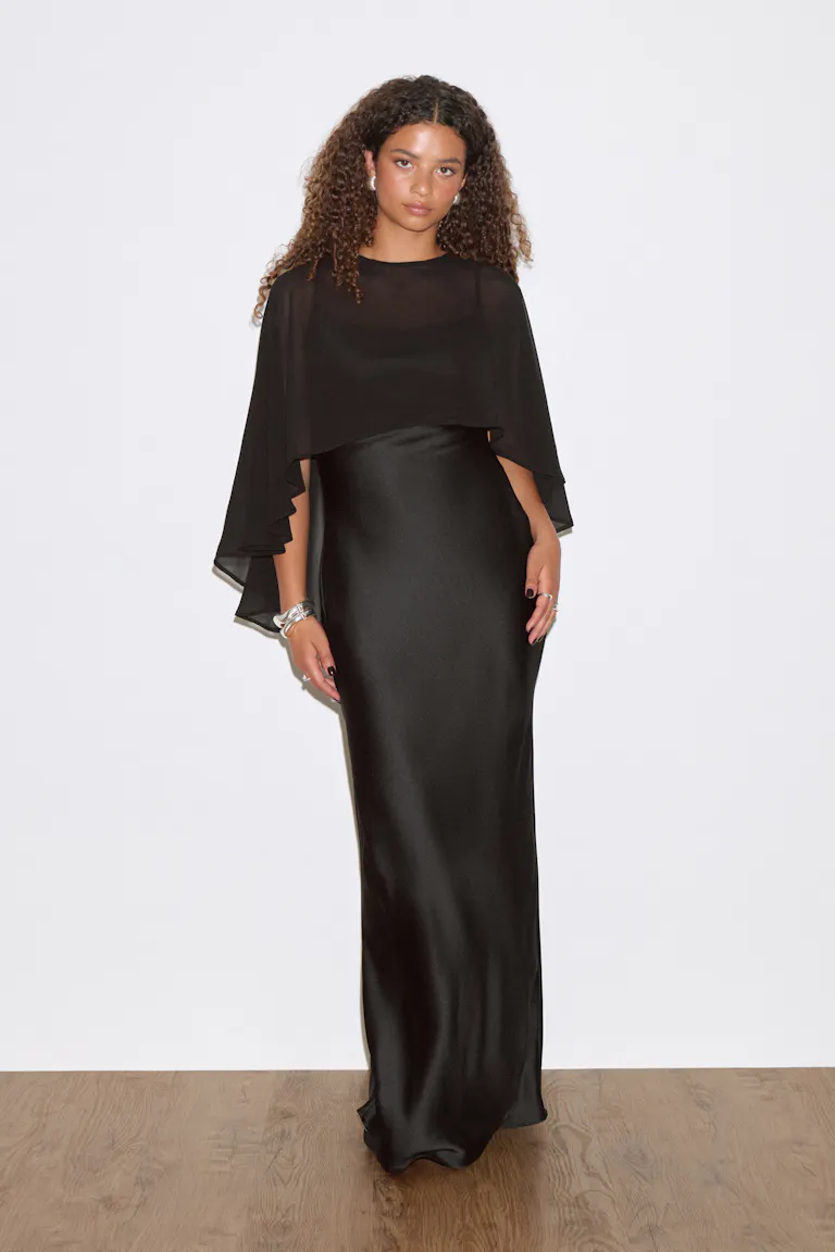 Black floor-length satin column gown with a round neckline and sheer chiffon cape overlay, styled with silver bracelets and rings.