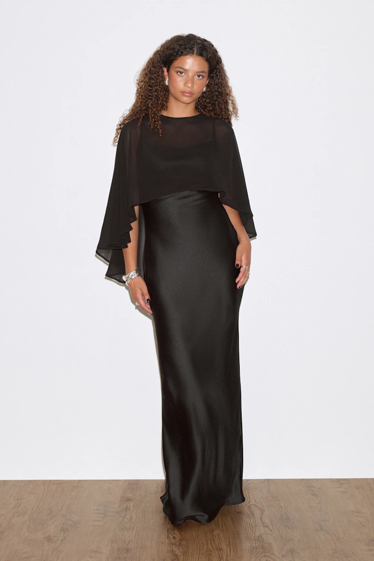 Zwarte zuiljurk van satijn met een ronde halslijn en een cape van doorschijnend chiffon, gestyled met zilveren armbanden en ringen.