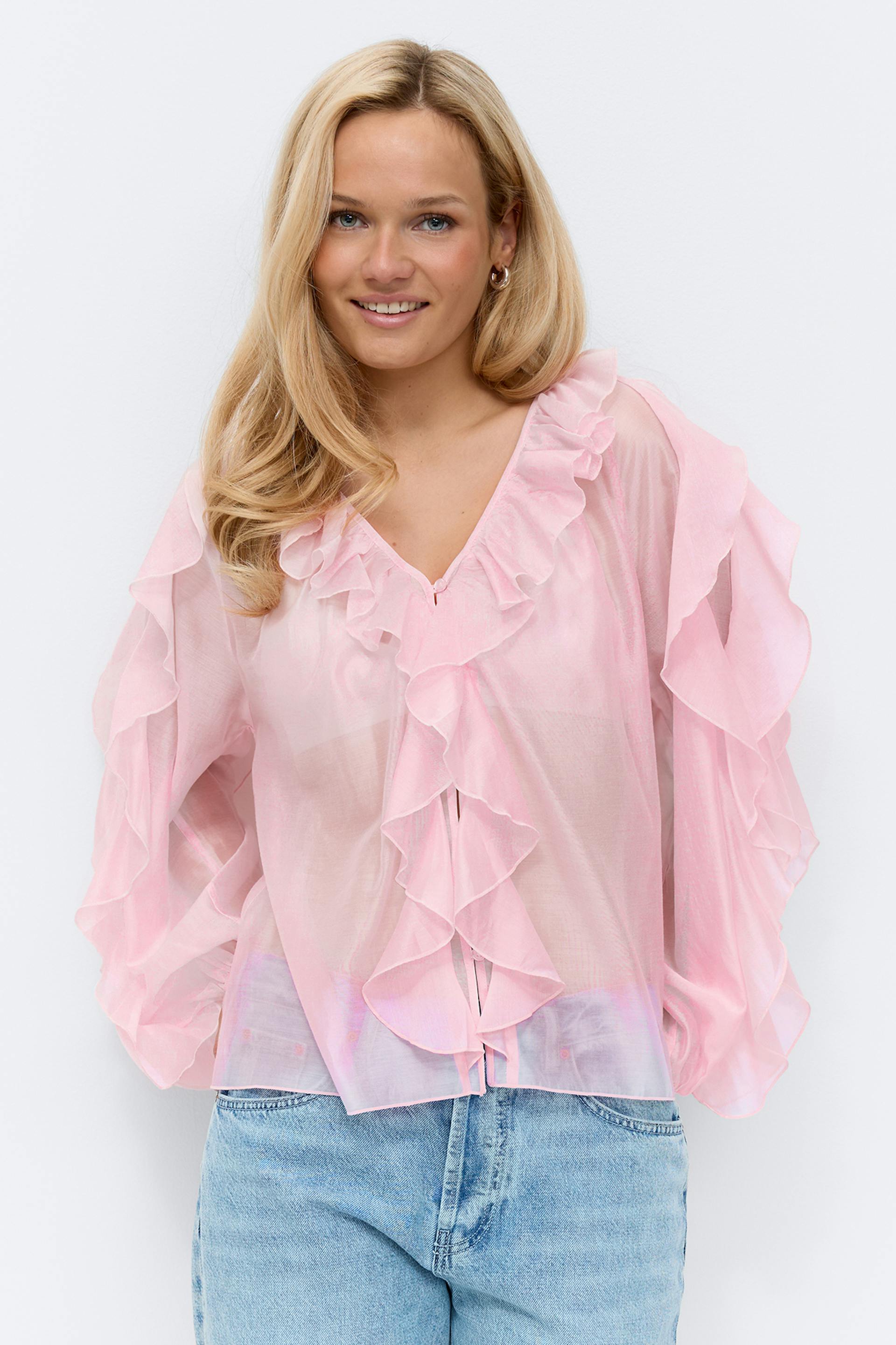 Pitkähihainen vaaleanvaaleanpunainen pusero, jossa on kerrostettuja röyhelöitä edessä ja hihoissa, yhdistettynä vaalean sinisiin farkkuihin.