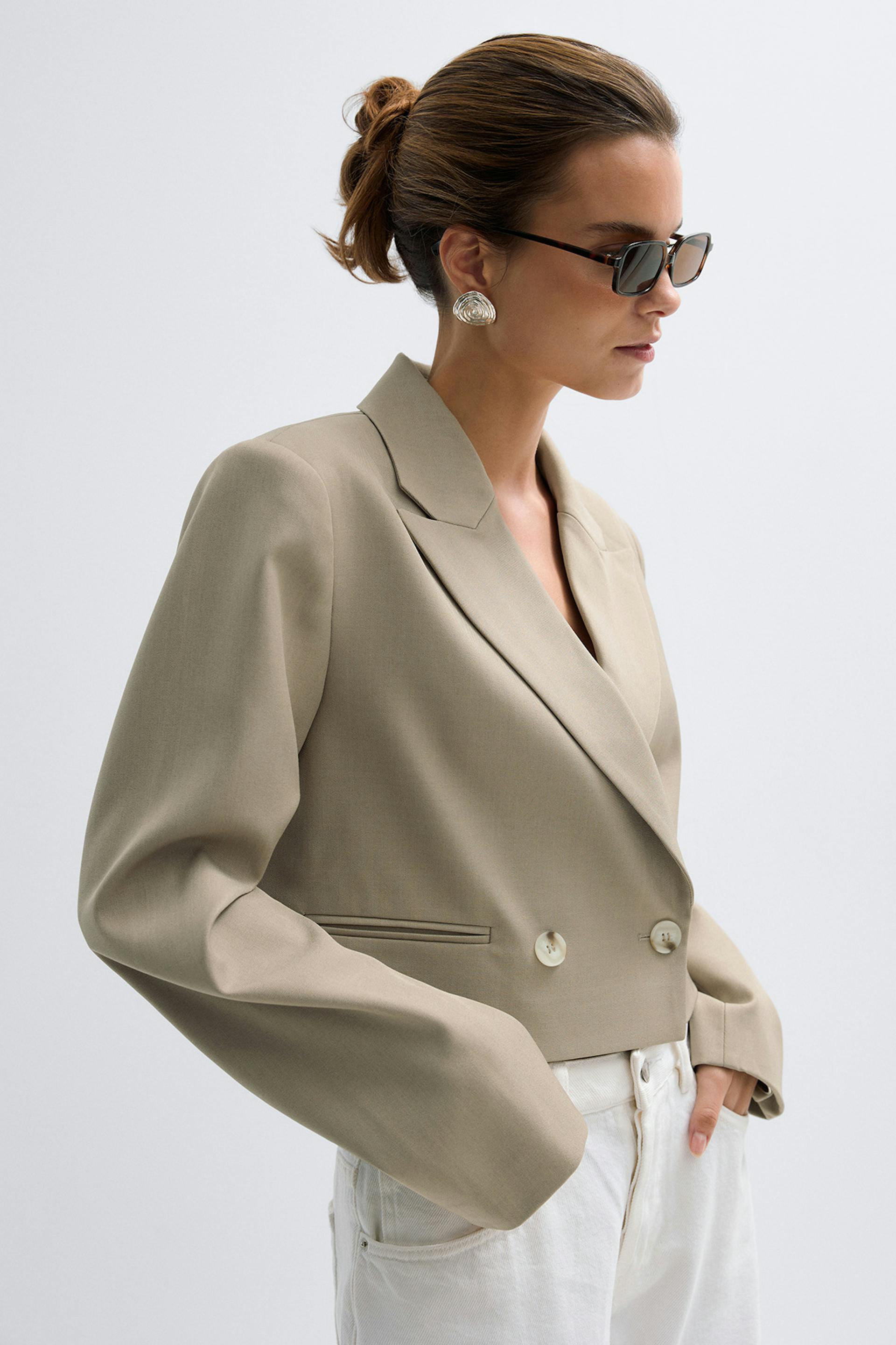 Blazer cropped beige à double boutonnage et à revers cranté, associé à un jean taille haute blanc, à des lunettes de soleil rectangulaires en écaille et à de grandes boucles d'oreilles argentées.