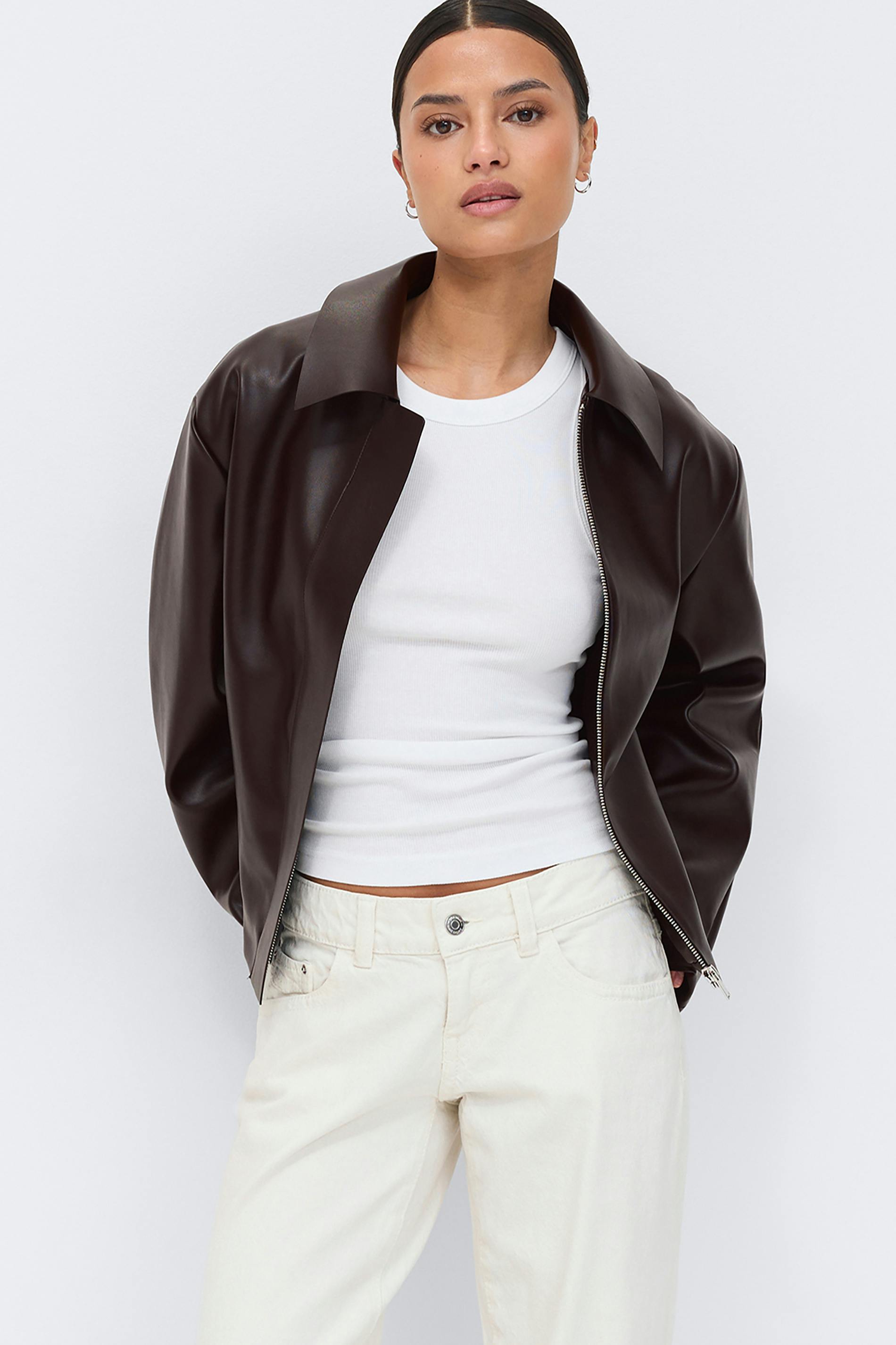 Dark brown collared leather zip-up jacket worn over a white ribbed sleeveless crew-neck top and cream straight-leg jeans.