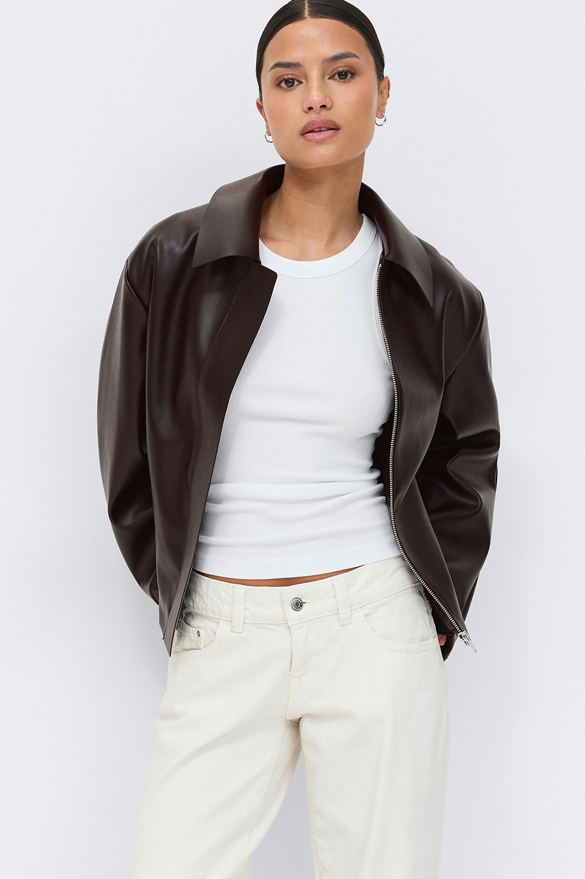 Dark brown collared leather zip-up jacket worn over a white ribbed sleeveless crew-neck top and cream straight-leg jeans.