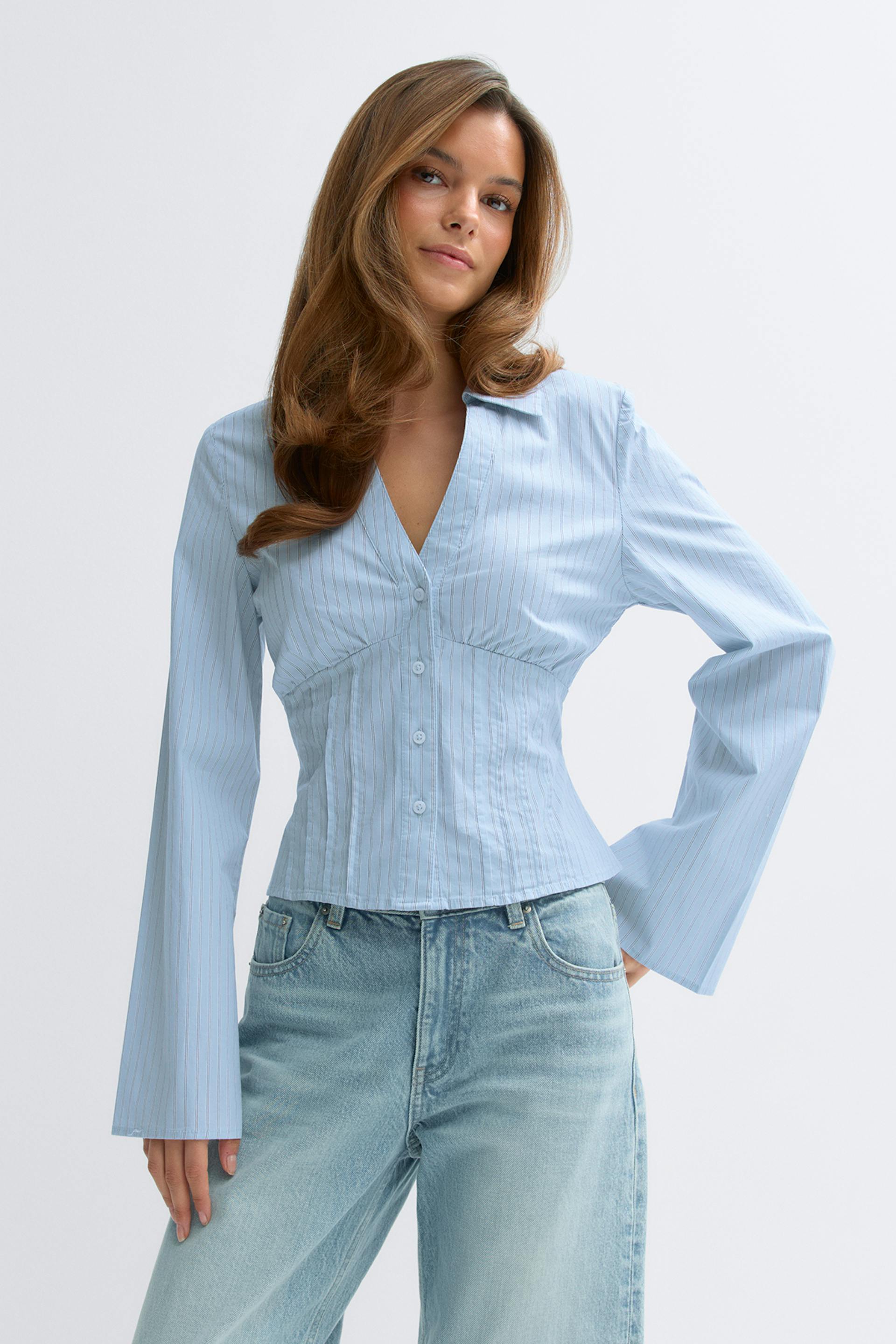 Hellblaue Nadelstreifenbluse mit Knopfleiste, V-Ausschnitt, taillierten/korsettierten Nähten und langen, ausgestellten Ärmeln, kombiniert mit hellen, hochgeschlossenen Jeans.