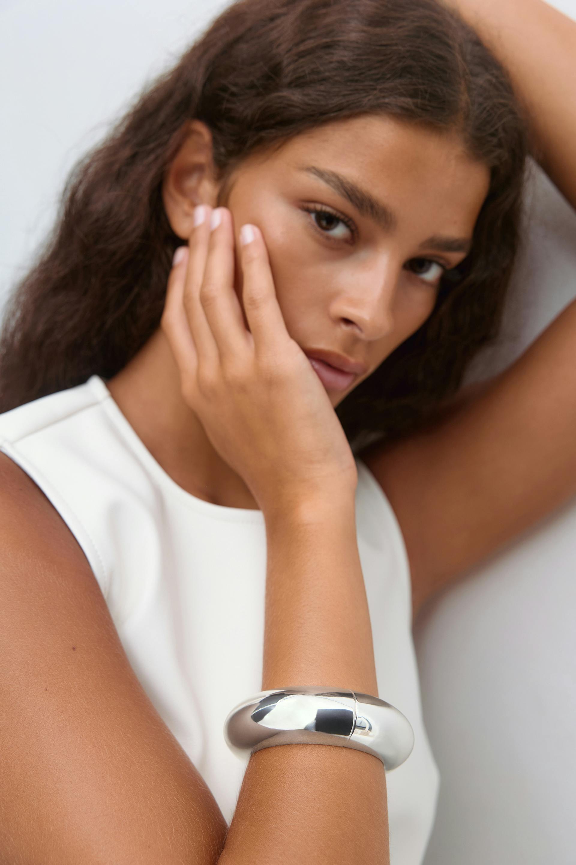 Sleeveless white crew‑neck top paired with a polished chunky silver cuff bracelet on the forearm.