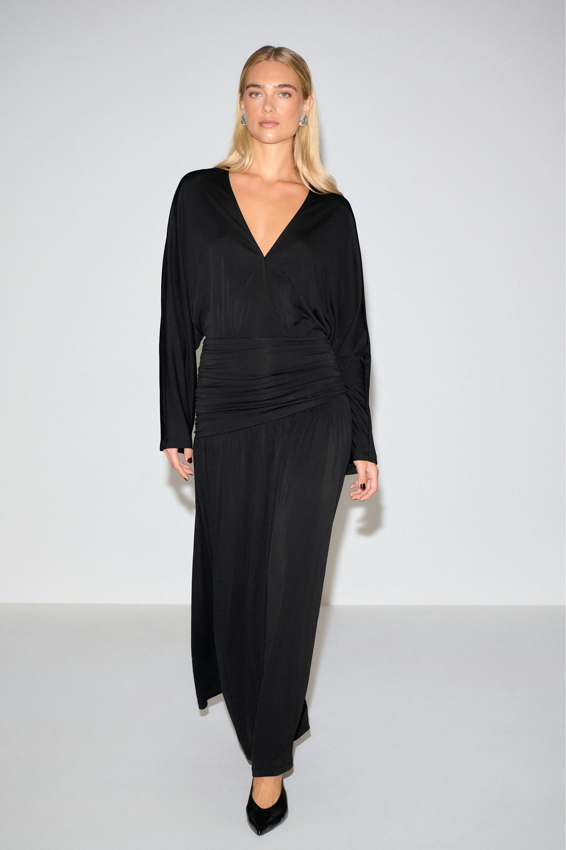 Black floor-length dress with a deep V-neck, long sleeves and a ruched waistband, paired with black pointed-toe heels.