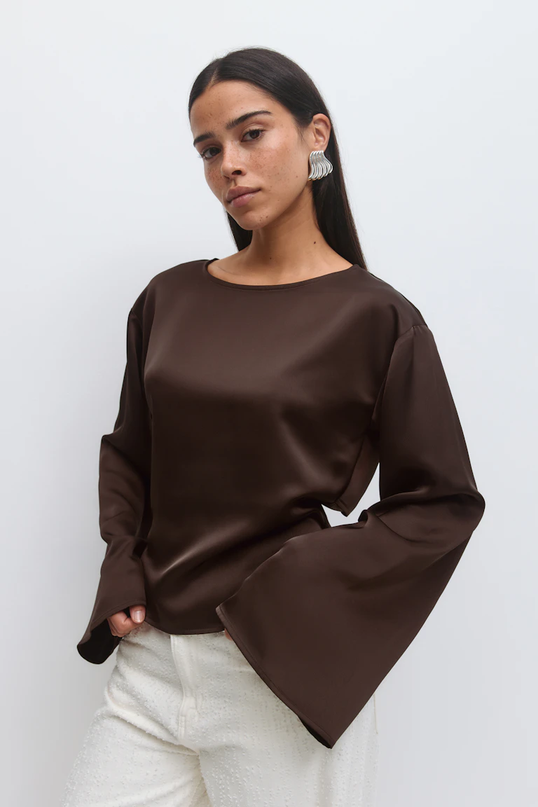 Blouse à manches longues en satin marron avec encolure ronde et manches cloche exagérées, associée à un pantalon texturé blanc et à des boucles d'oreilles ondulées argentées.