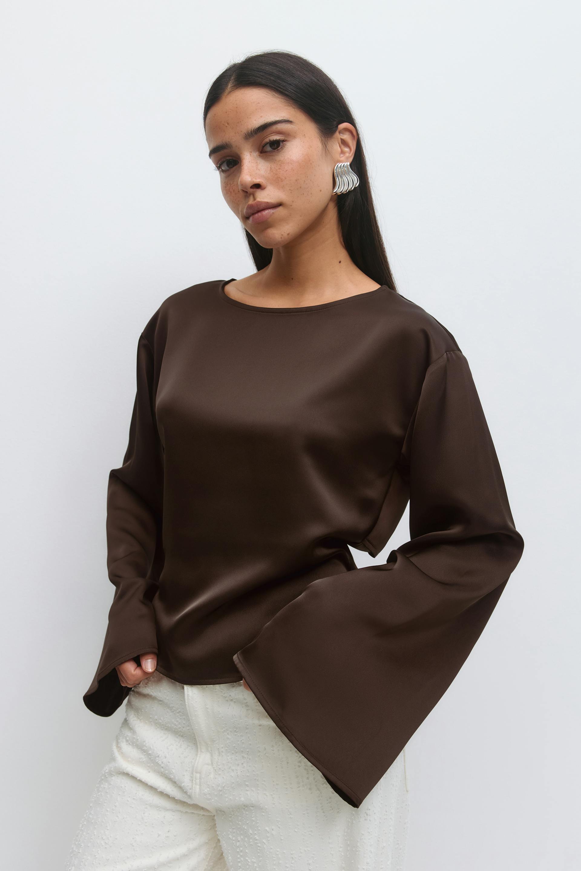 Bruine satijnen blouse met lange mouwen, ronde hals en overdreven klokmouwen, gecombineerd met een witte broek met structuur en zilveren, golvende oorbellen.