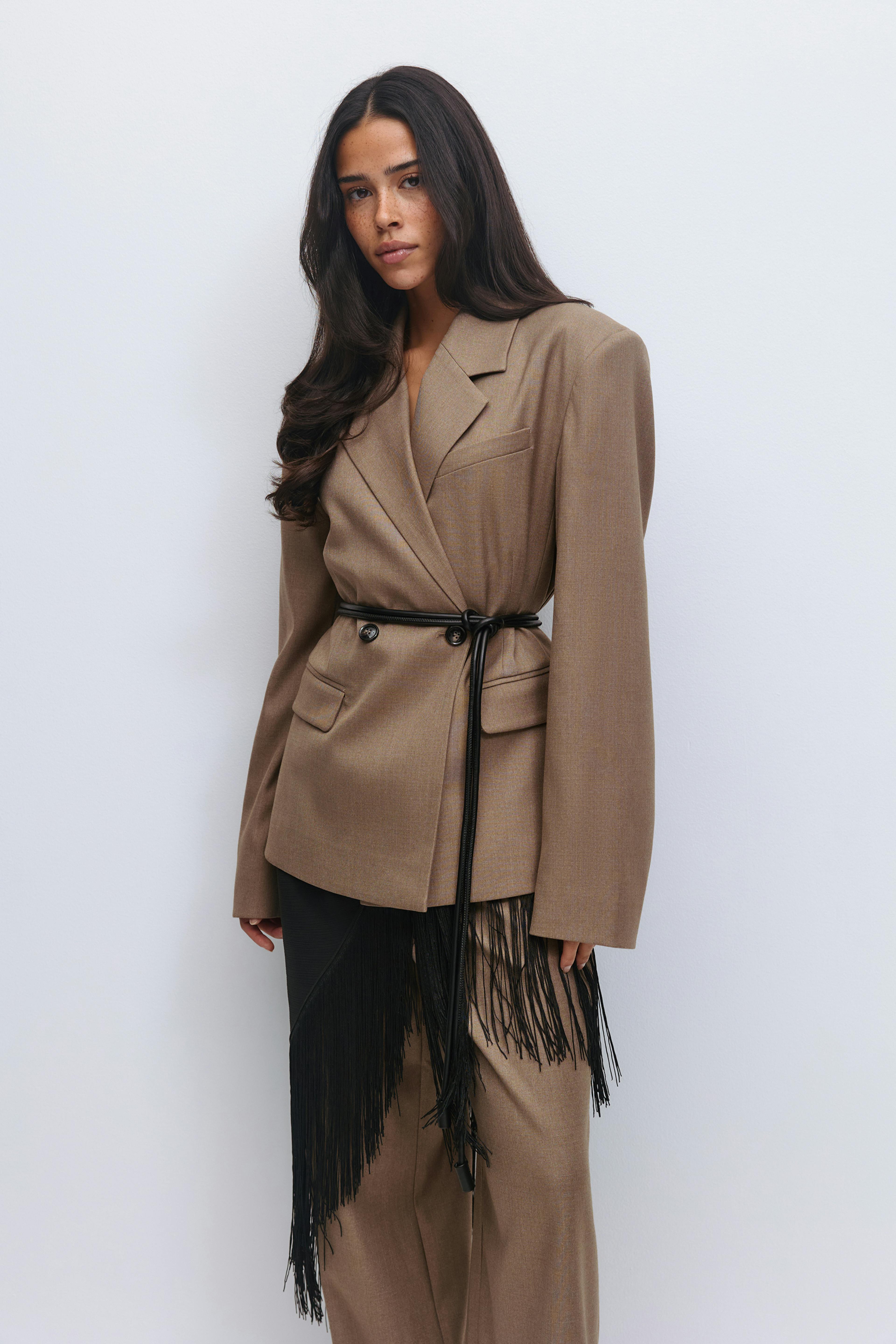 Taupekleurige getailleerde blazer met dubbele rij knopen en klep- en borstzakken, vastgemaakt met een dunne zwarte ceintuur, gecombineerd met een bijpassende broek met wijde pijpen en een asymmetrisch paneel met zwarte franjes.