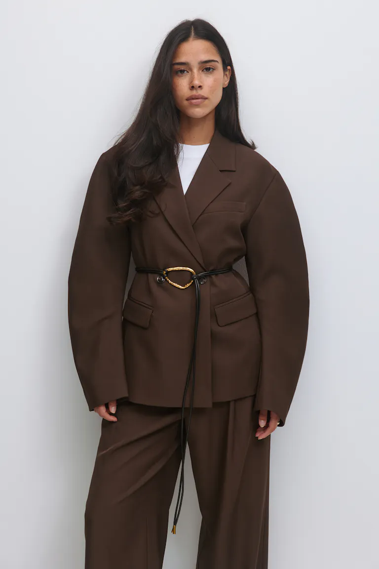 Schokoladenbrauner, übergroßer, maßgeschneiderter Blazer, der in der Taille mit einer dünnen schwarzen Lederkordel mit unregelmäßiger ovaler Schnalle in Gold gegürtet wird, kombiniert mit einer passenden Hose mit weitem Bein und einem weißen Oberteil mit Rundhalsausschnitt.