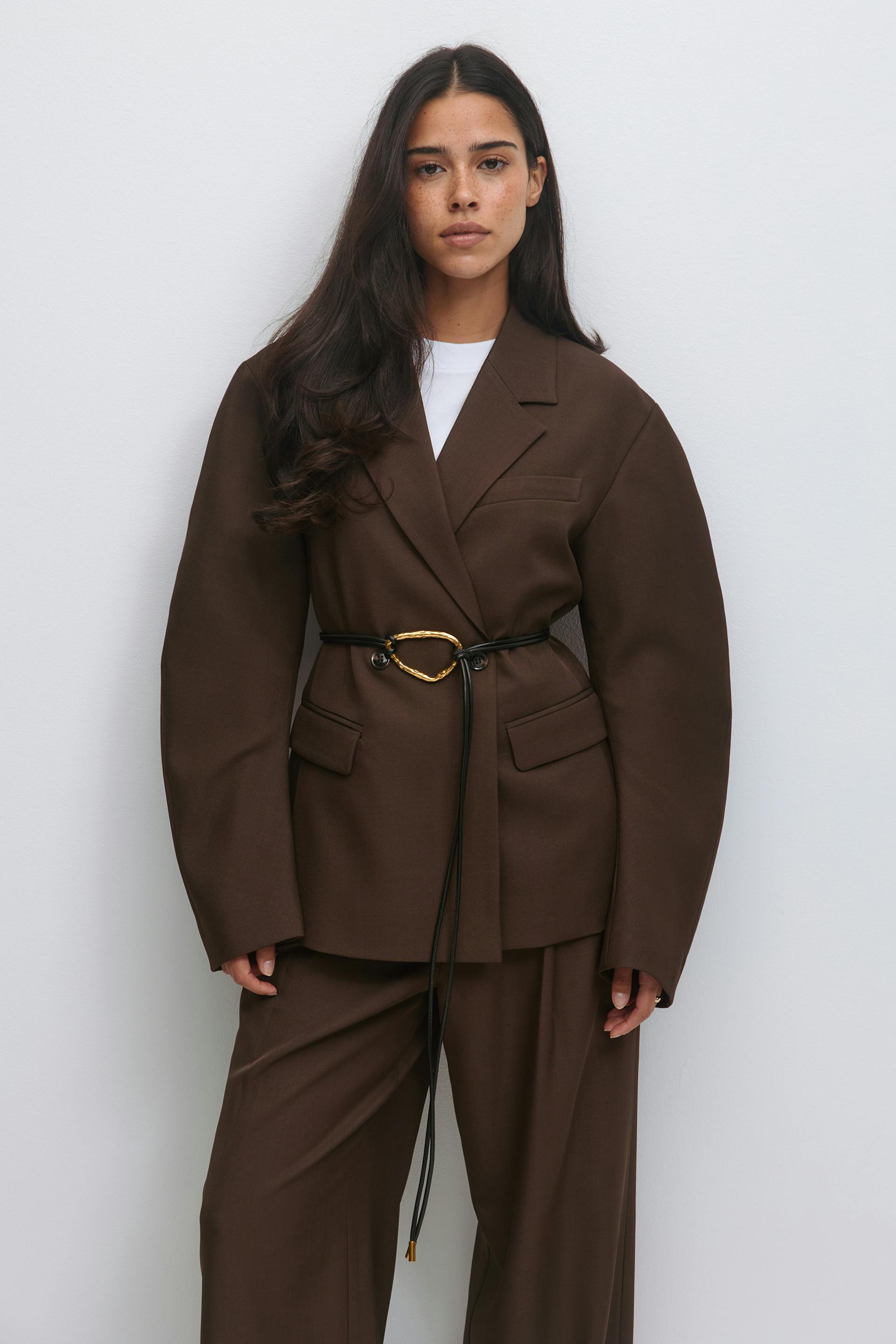 Chocolate-brown oversized tailored blazer belted at the waist with a thin black leather cord featuring a gold irregular oval buckle, paired with matching wide-leg trousers and a white crew-neck top.