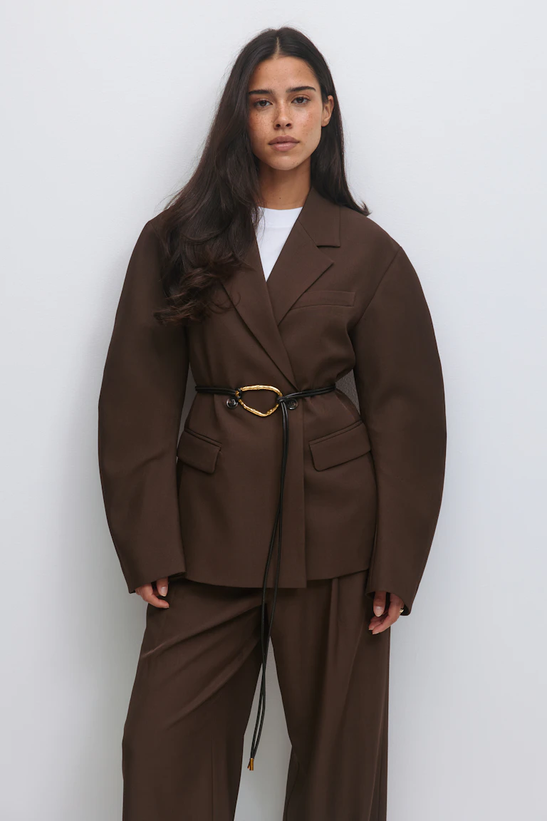 Chocoladebruine oversized getailleerde blazer die in de taille is vastgemaakt met een dun zwart leren koord met een gouden onregelmatige ovale gesp, gecombineerd met een bijpassende broek met wijde pijpen en een witte top met ronde hals.