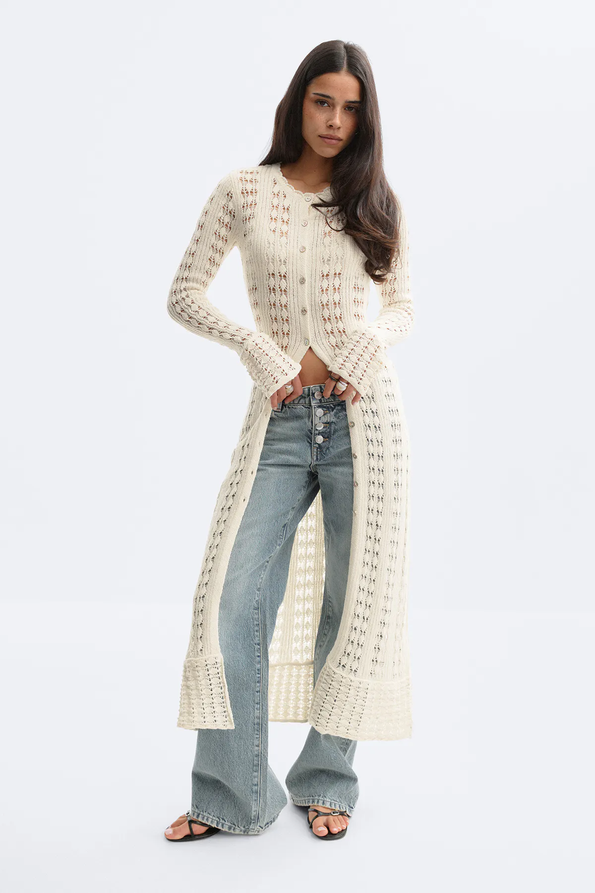 Pitkä kermanvärinen virkattu neulepusero, jossa on kauluri kaula-aukko ja levenevät hihansuut, yhdistettynä korkeavyötäröisiin vaaleisiin farkkuihin ja mustiin remmikorokkeellisiin sandaaliin.