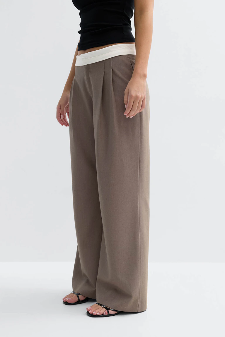 Top cropped sans manches noir associé à un pantalon large taupe à taille haute, avec plis sur le devant et ceinture blanche repliée ; complété par des sandales plates noires à lanières.