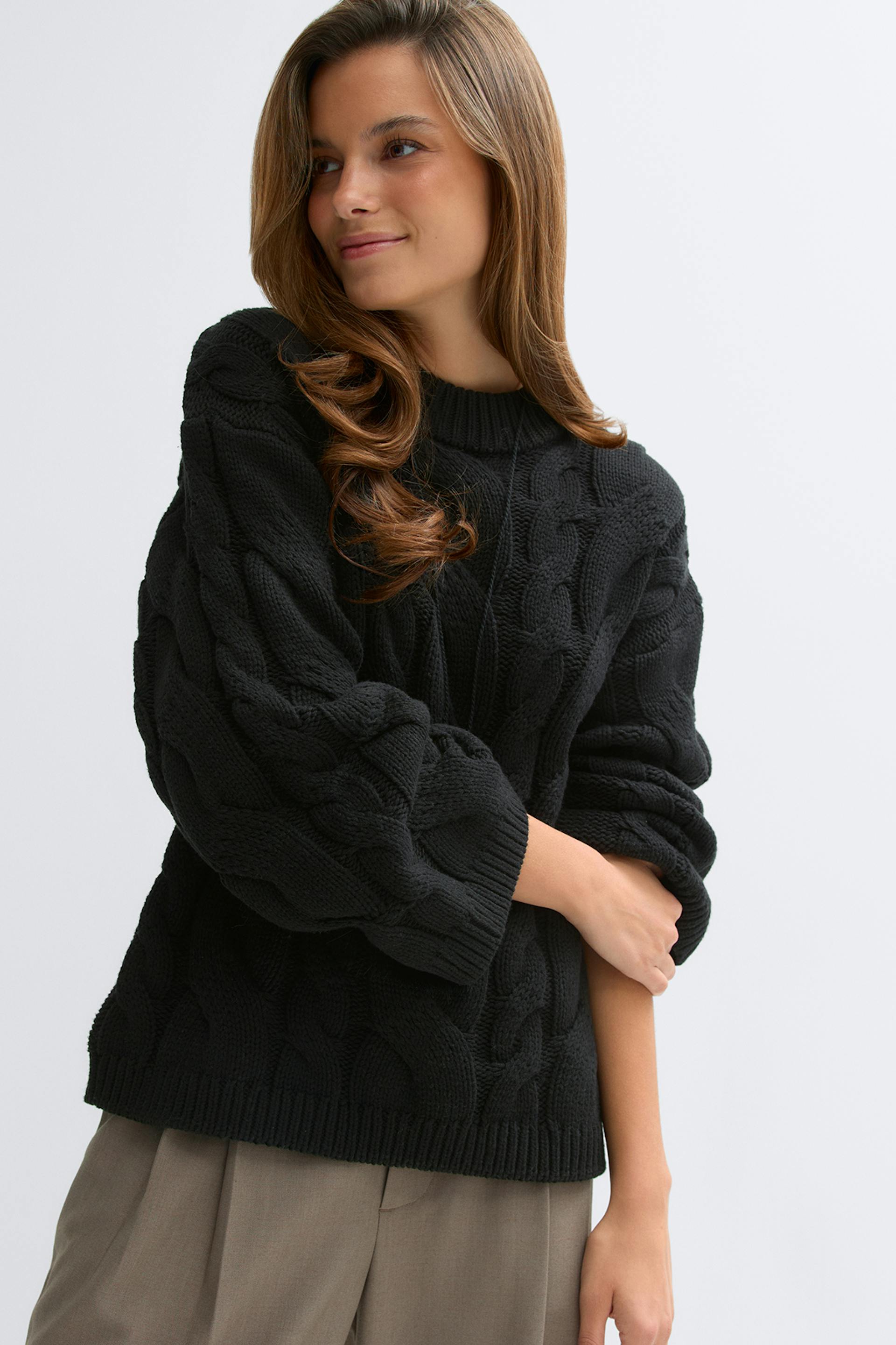 Schwarzer Grobstrickpullover mit Rundhalsausschnitt, Ballonärmeln und gerippten Bündchen und Saum, kombiniert mit einer taupefarbenen Bundfaltenhose.