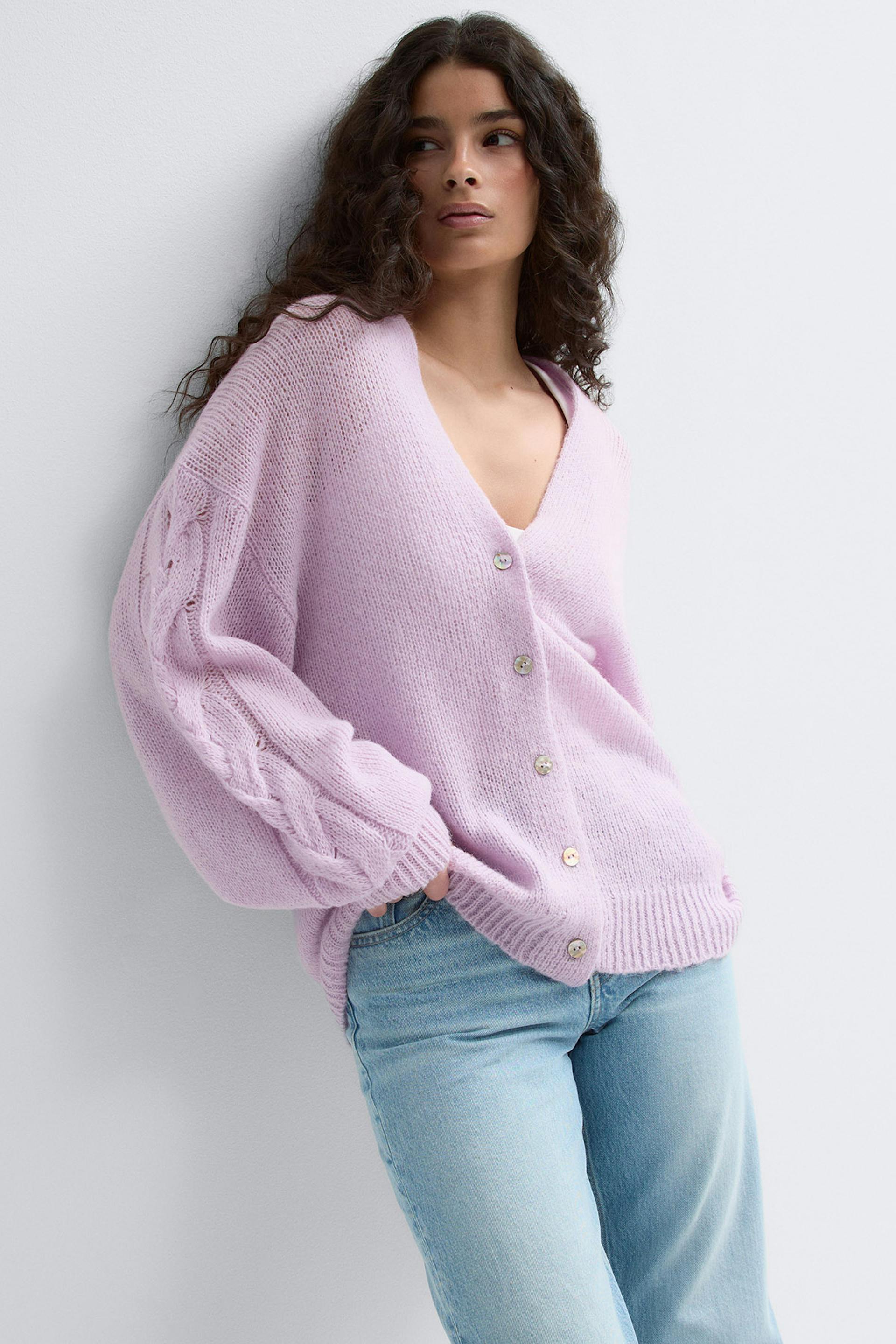 Heller lavendelfarbener Strickcardigan mit V-Ausschnitt und Knopfleiste in lockerer Passform und geflochtenen Zopfdetails an den Ballonärmeln, kombiniert mit hellen Jeans mit geradem Bein.