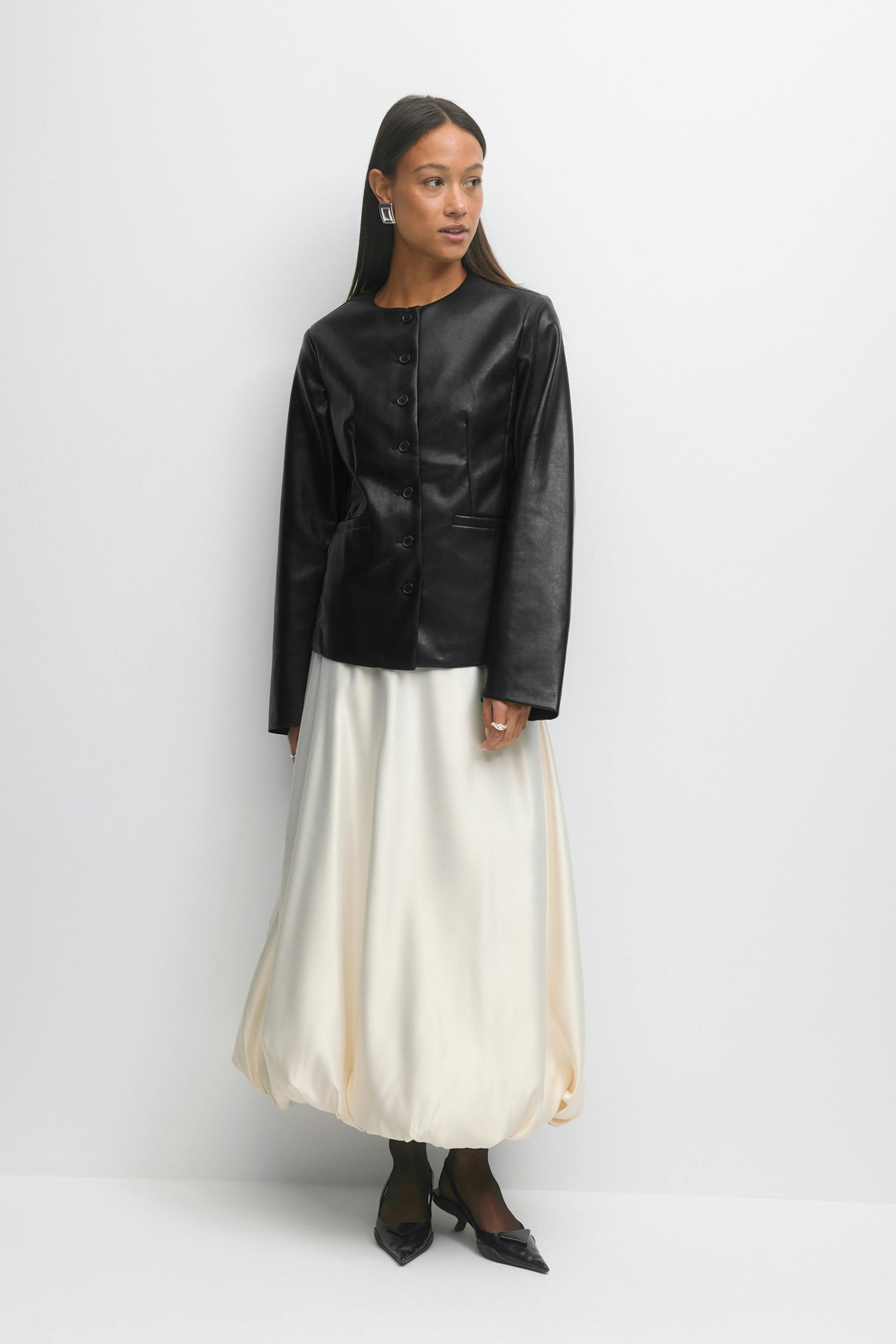 Musta pitkähihainen nahkatakki, jossa on nappikiinnitys, norsunluunvärinen satiinipallohelma, mustat läpikuultavat sukkahousut, teräväkärkiset mustat matalakorkoiset korkokengät ja geometriset hopeiset korvakorut.