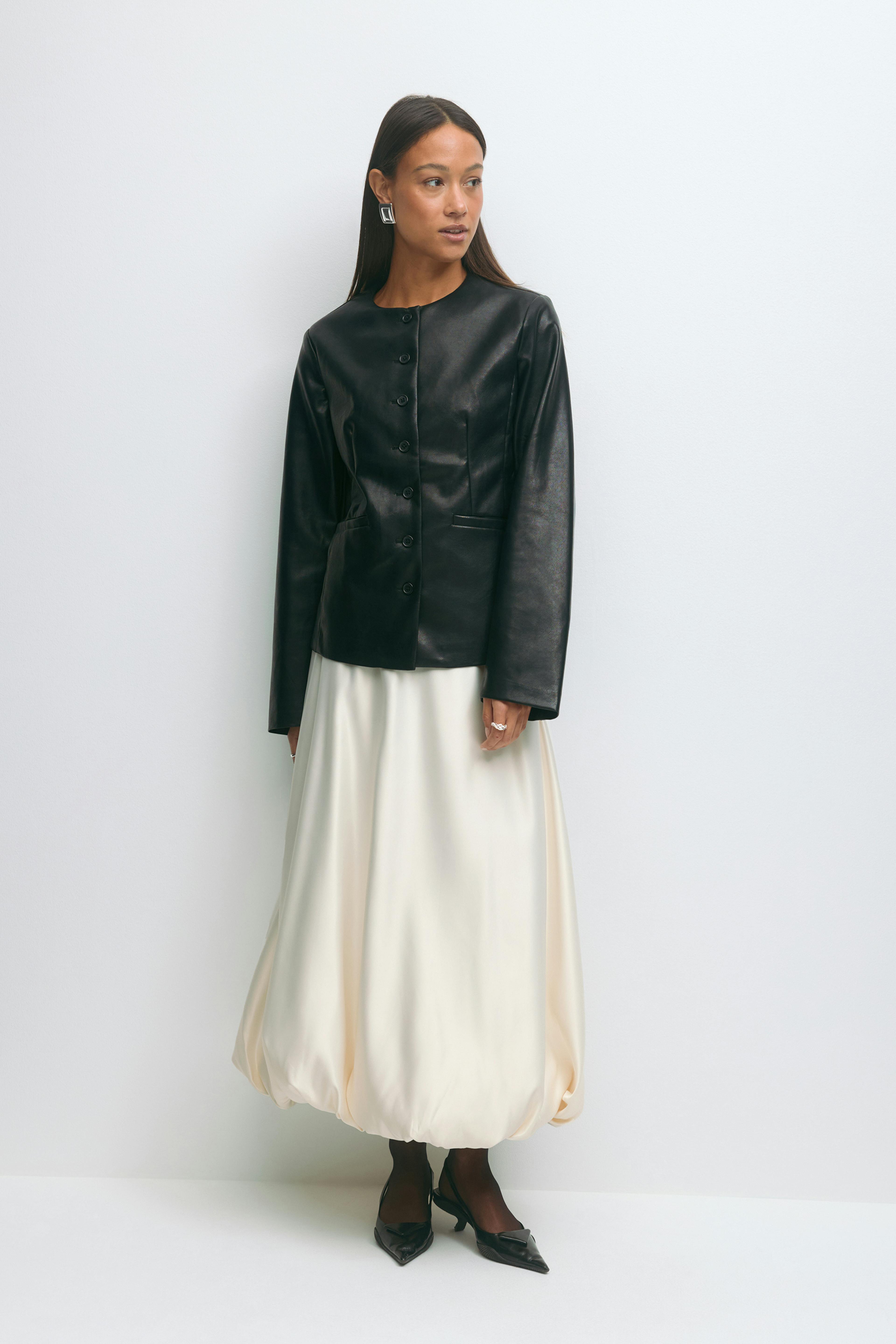 Zwart leren jasje met lange mouwen en knoopsluiting, ivoorkleurige satijnen midi-rok met ballonzoom, zwarte doorschijnende panty, spitse zwarte pumps met lage hak en geometrische zilveren oorbellen.