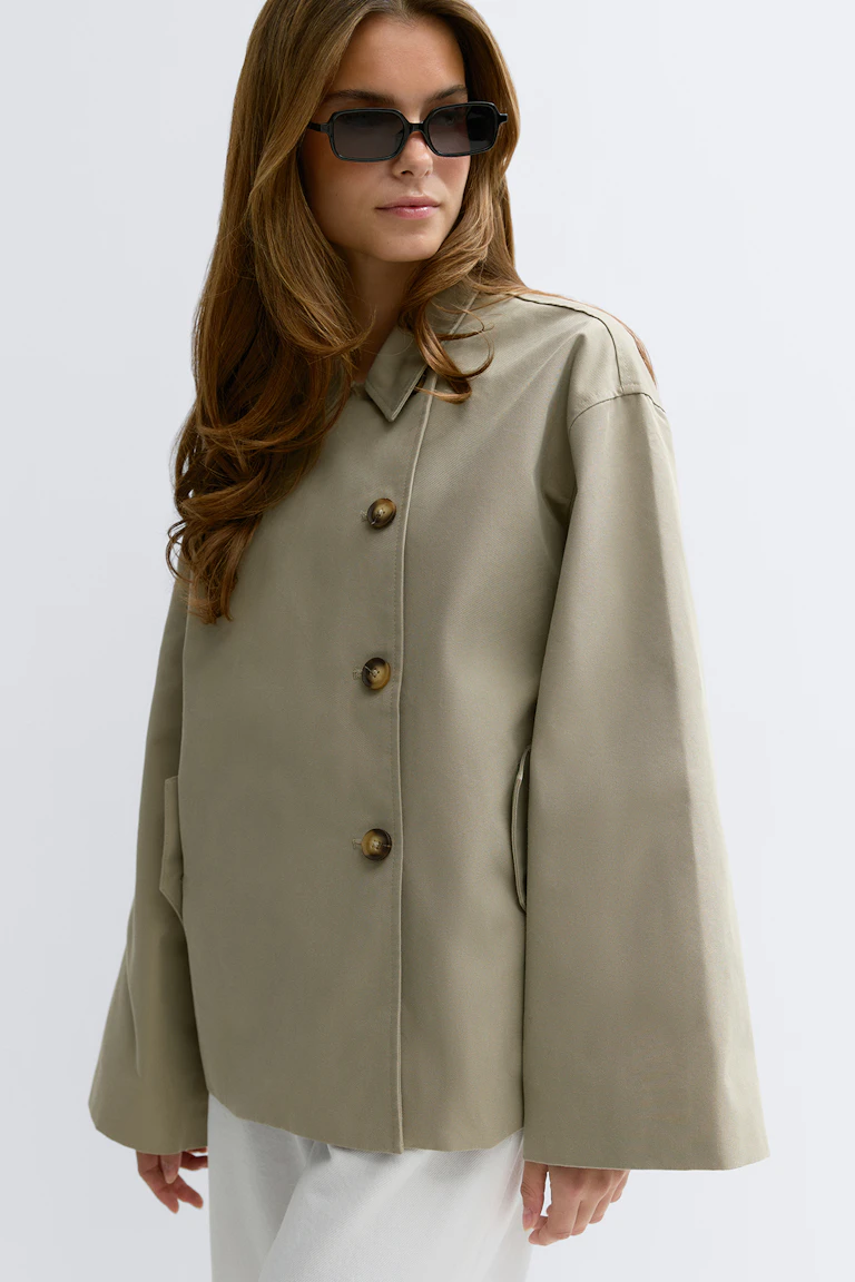 Light khaki oversized button-front jacket with wide sleeves and notch collar, paired with black rectangular sunglasses and white trousers.