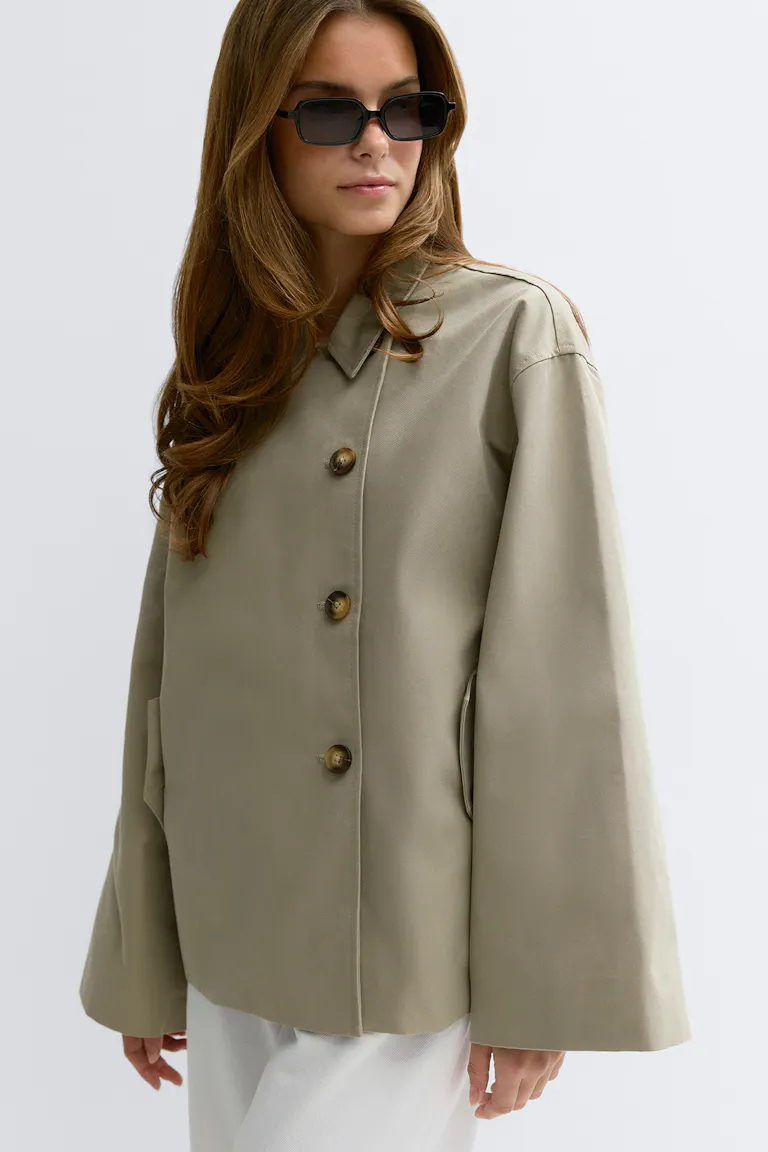 Helle khakifarbene Oversize-Knopfleiste mit weiten Ärmeln und Kragen, gepaart mit einer schwarzen rechteckigen Sonnenbrille und einer weißen Hose.