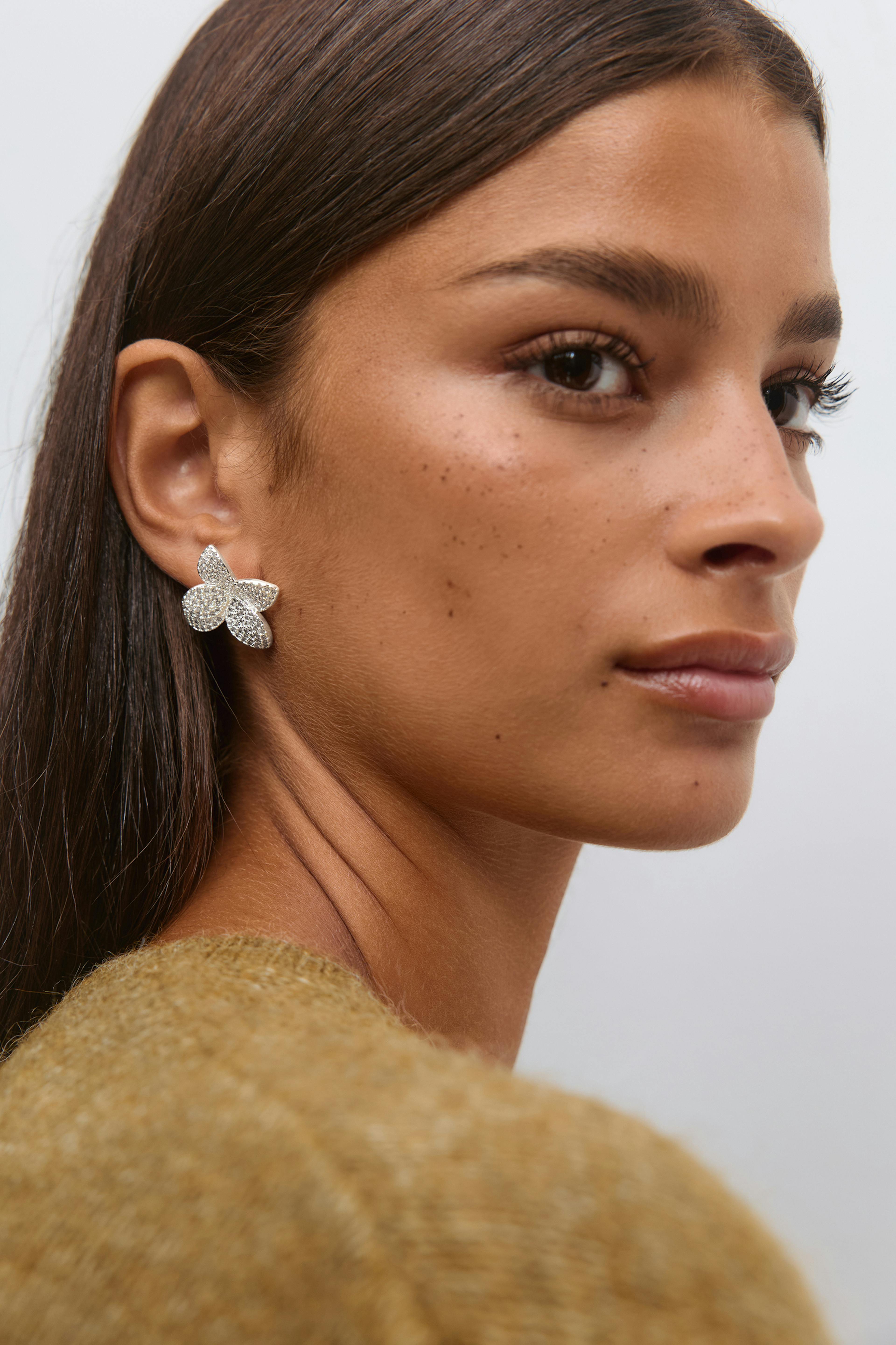 Modellen bär silverfärgade, kristallinfattade blomörhängen och en senapsfärgad, fluffig stickad tröja.