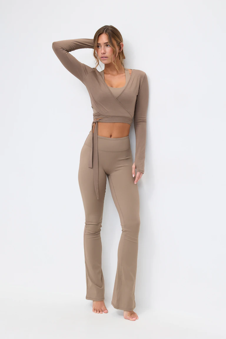 Taupefarbenes langärmeliges Wickel-Crop-Top mit seitlichem Bindeband über einem passenden Sport-BH mit Rundhalsausschnitt, gepaart mit hochtaillierten, ausgestellten Leggings in der Farbe Taupe.
