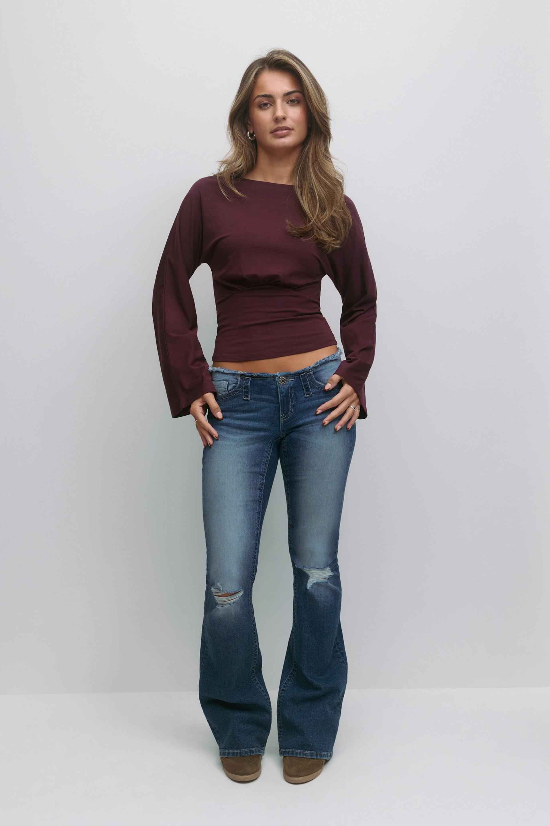 Long-sleeve maroon fitted cropped top with a ruched waist, paired with blue distressed flared jeans featuring knee rips and brown suede ankle boots.