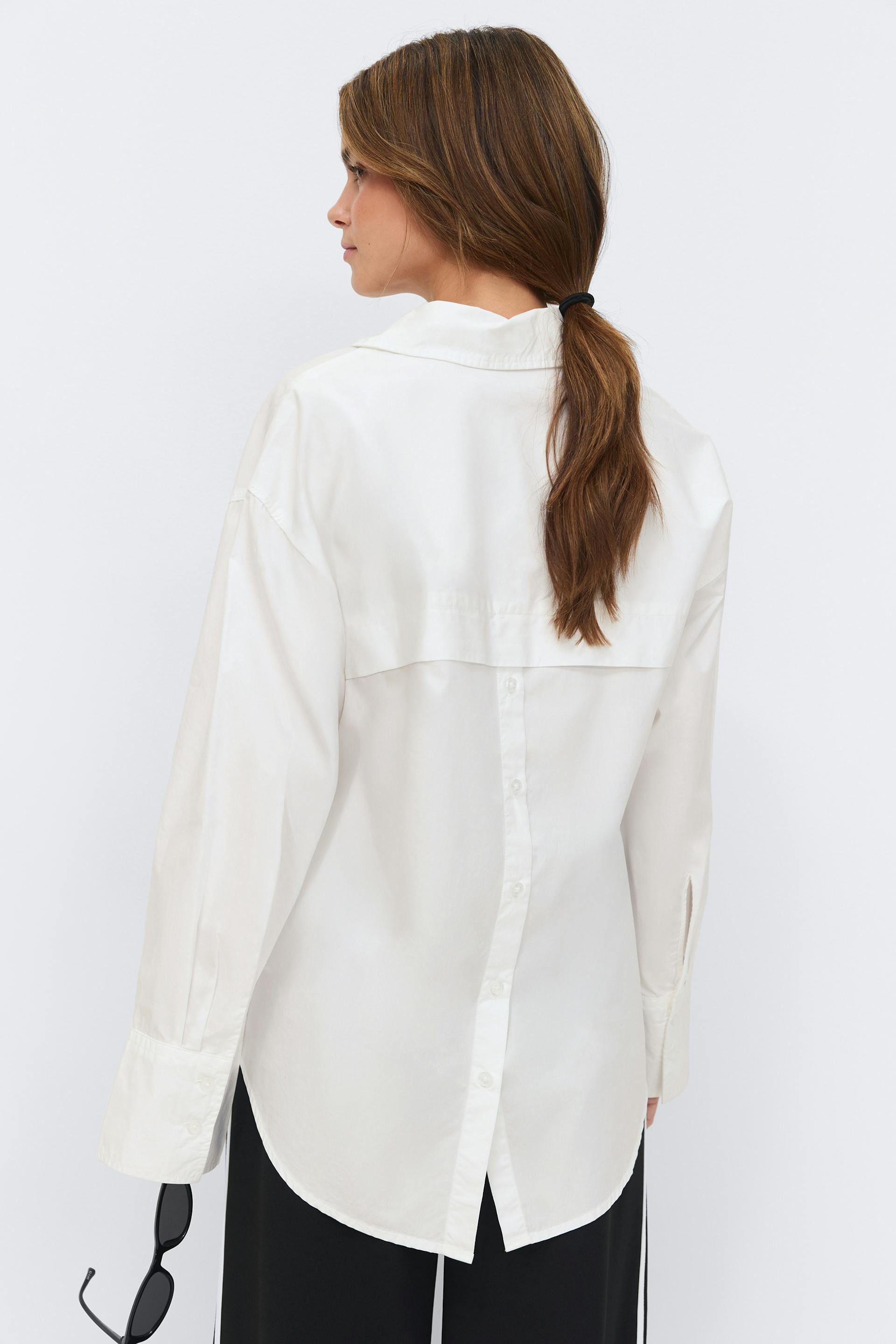 White long-sleeve collared blouse with button-back detailing, extended cuffs and curved hem, paired with black trousers; model holding black sunglasses.