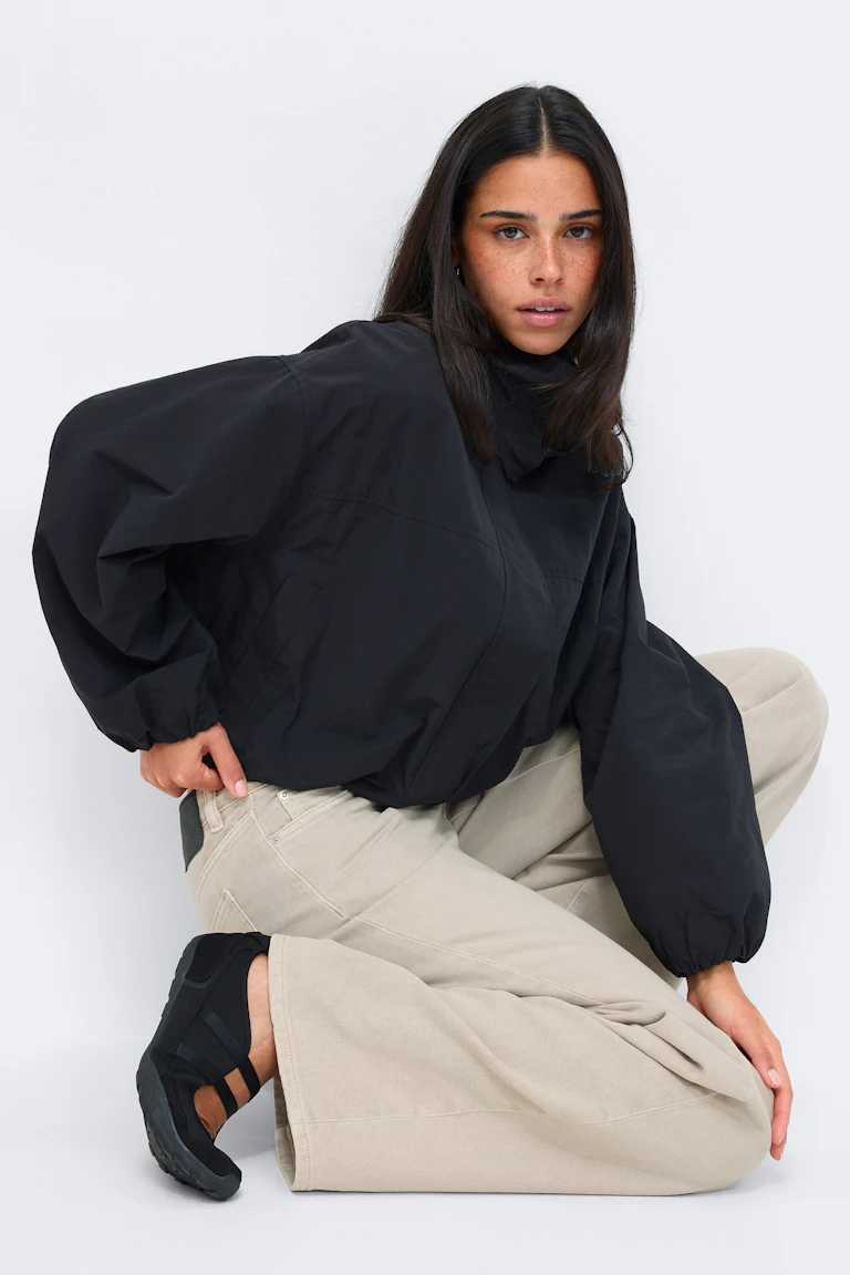 Schwarze Oversize-Jacke mit hohem Kragen und elastischen Bündchen, kombiniert mit einer beigen Hose mit weitem Bein und schwarzen Slip-on-Sneakern mit elastischen Riemen.