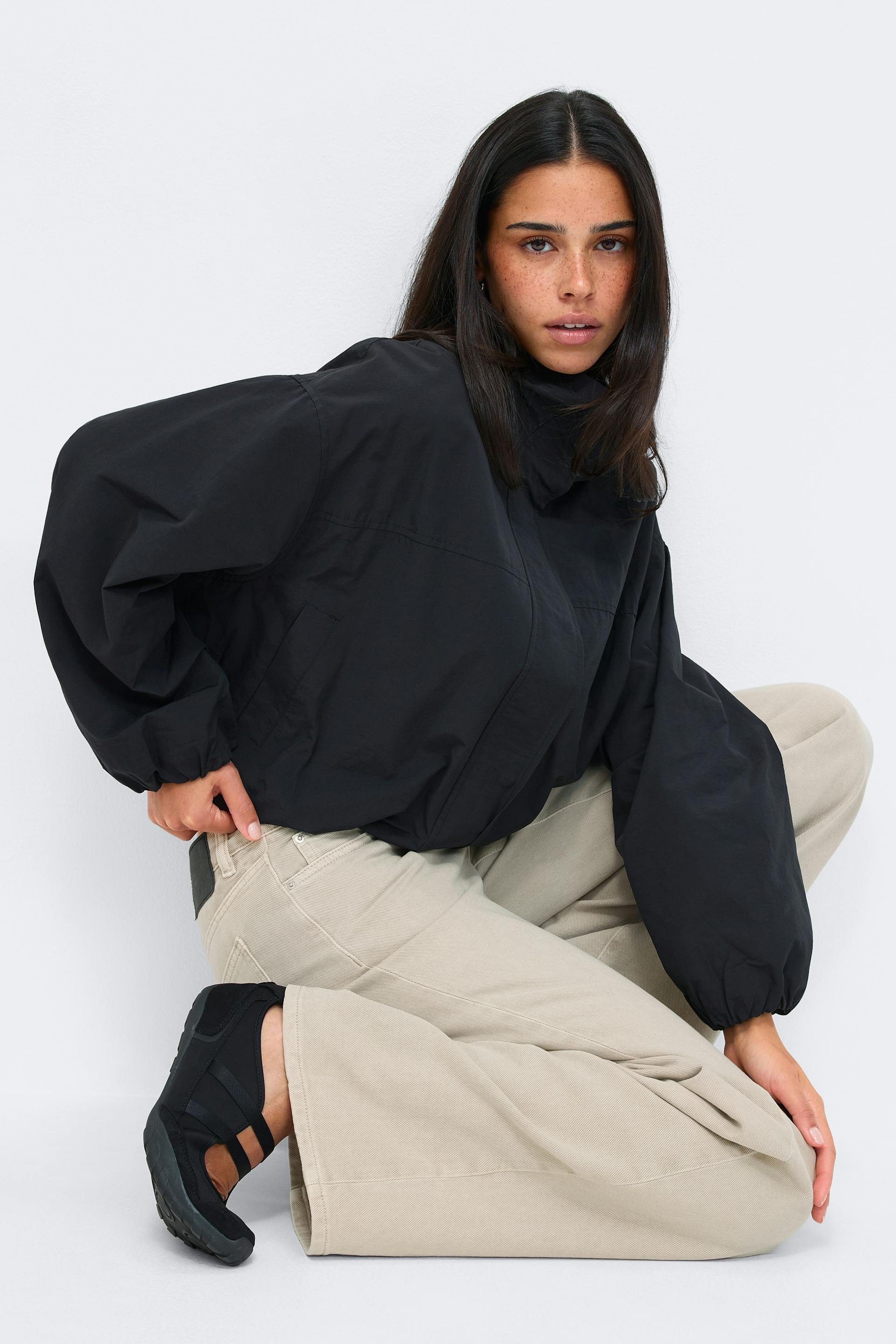 Black oversized high‑collar jacket with elasticized cuffs, paired with beige wide‑leg trousers and black slip‑on sneakers with elastic straps.