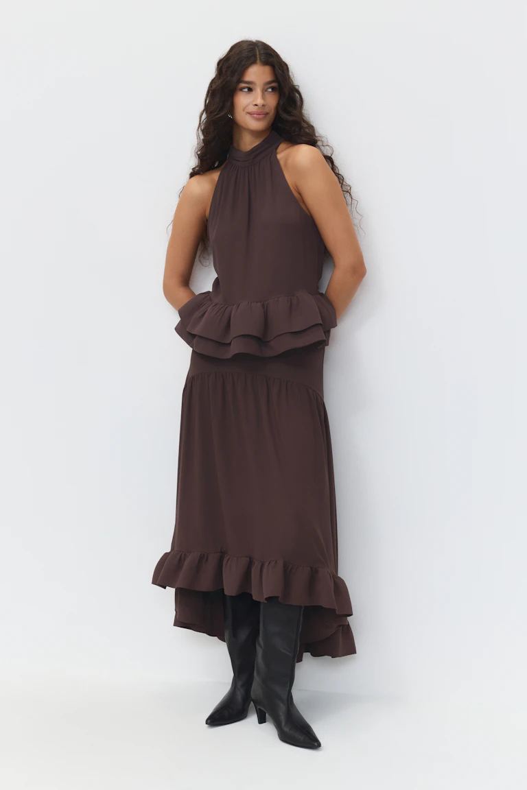 Ärmelloses, hochgeschlossenes, dunkelbraunes Midikleid mit gestuftem Schößchen und gerüschtem High-Low-Saum, kombiniert mit schwarzen, kniehohen, spitz zulaufenden Stiefeln.