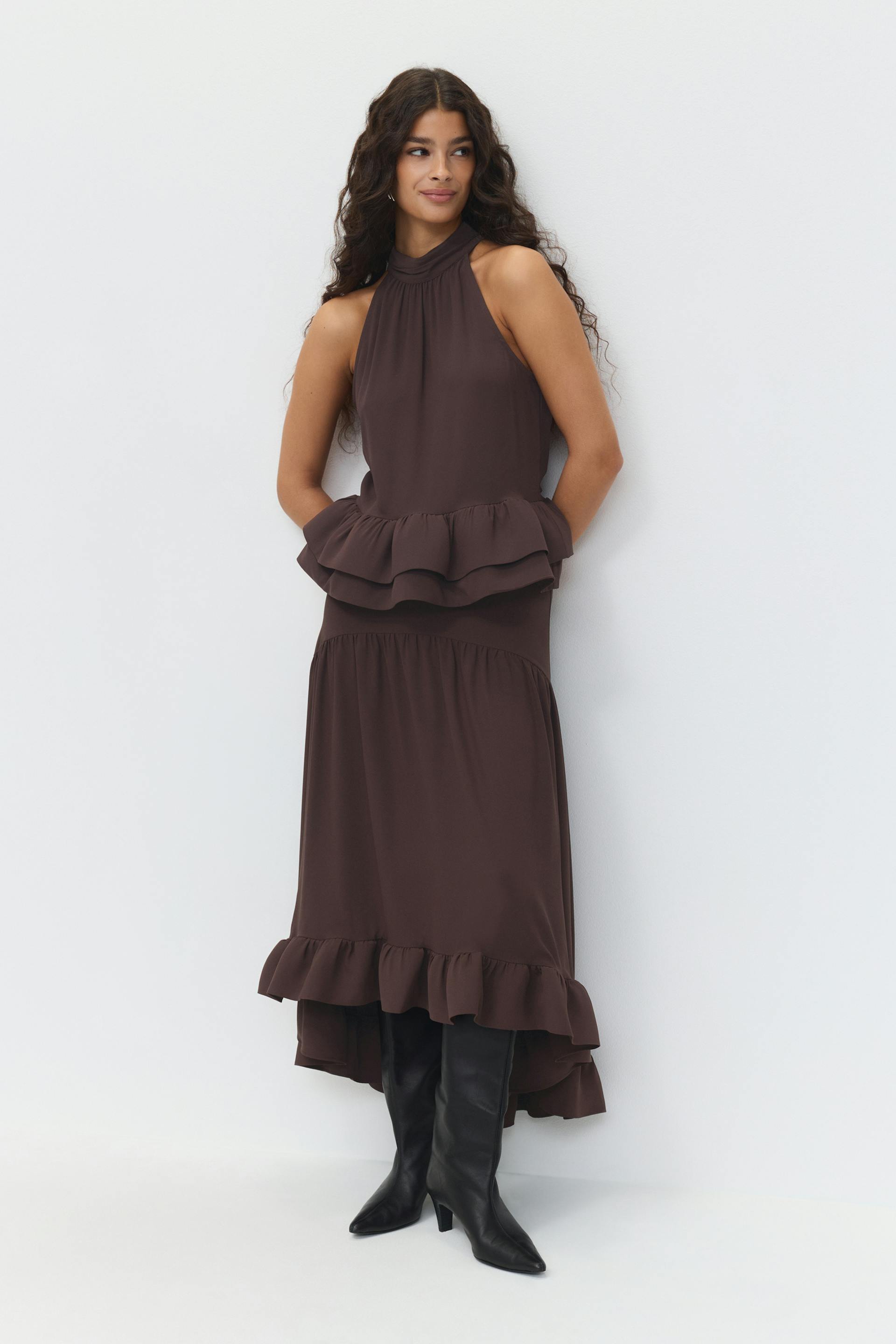 Sleeveless high‑neck deep‑brown midi dress with a tiered peplum and ruffled high‑low hem, paired with black knee‑high pointed‑toe heeled boots.