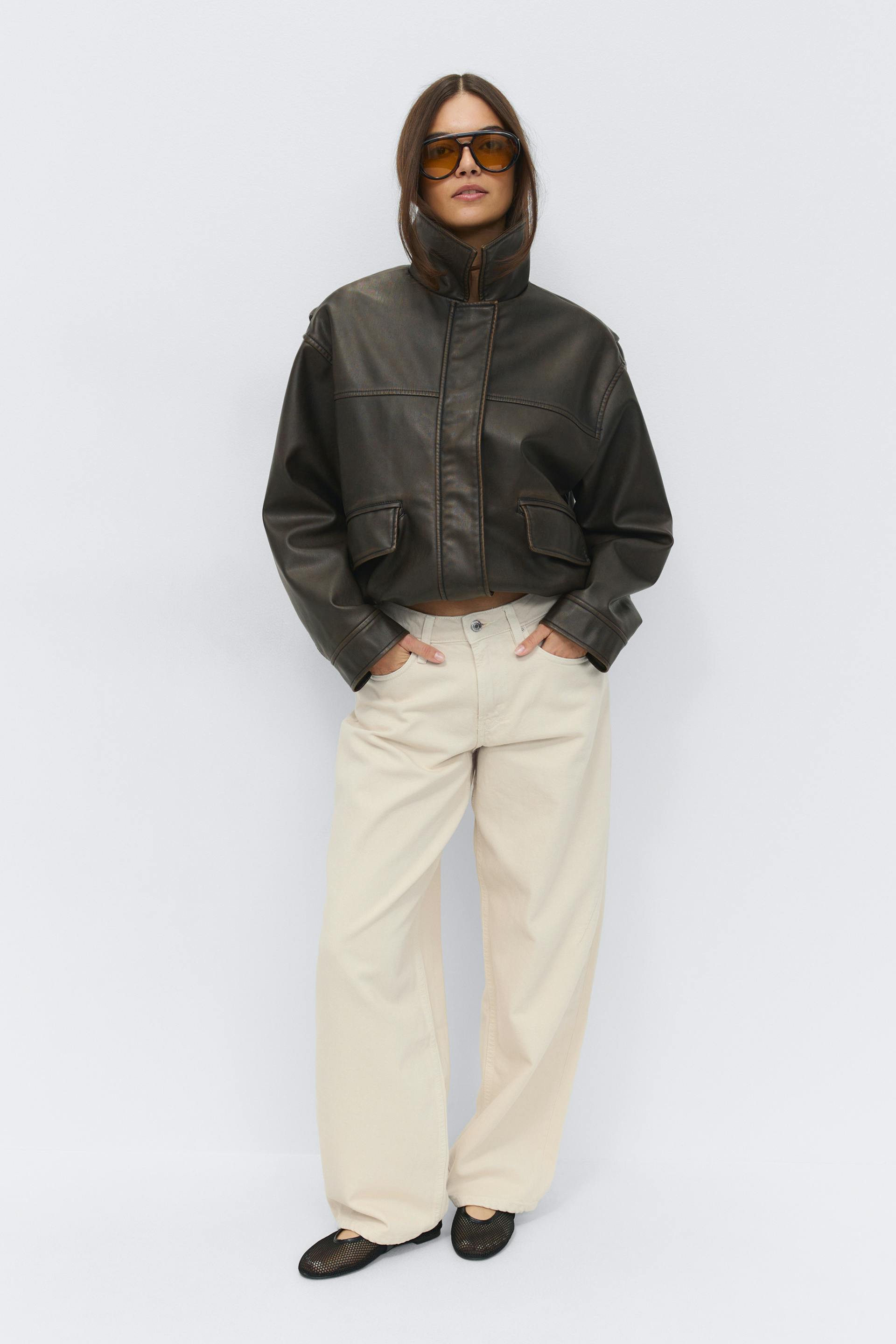 Model wearing a dark brown cropped leather jacket with high collar and flap pockets, cream wide-leg trousers, black mesh ballet flats and oversized amber-tinted sunglasses.