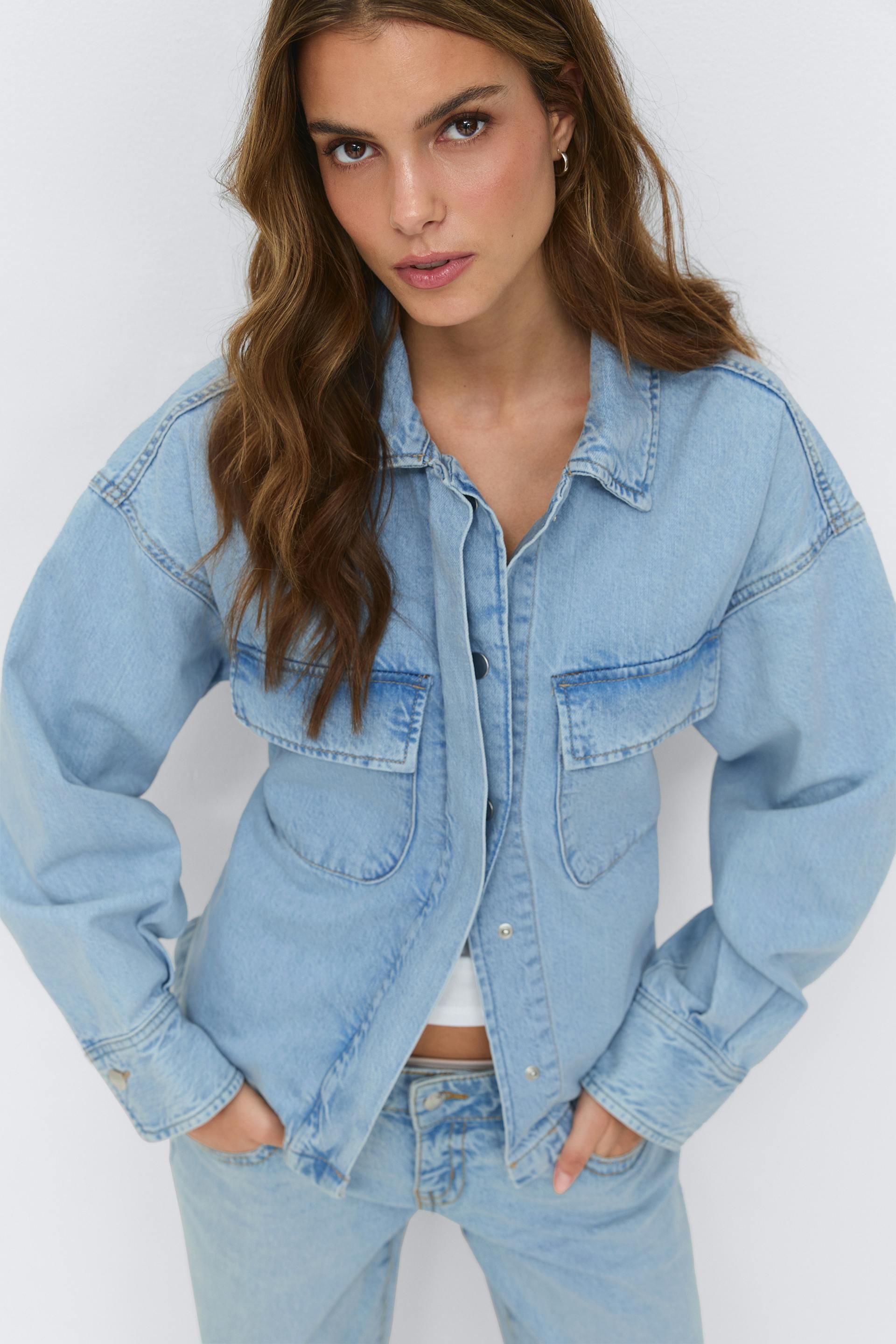 Light-wash denim jacket with flap chest pockets and snap-front closure, worn over a white cropped top and paired with matching light-wash denim jeans.