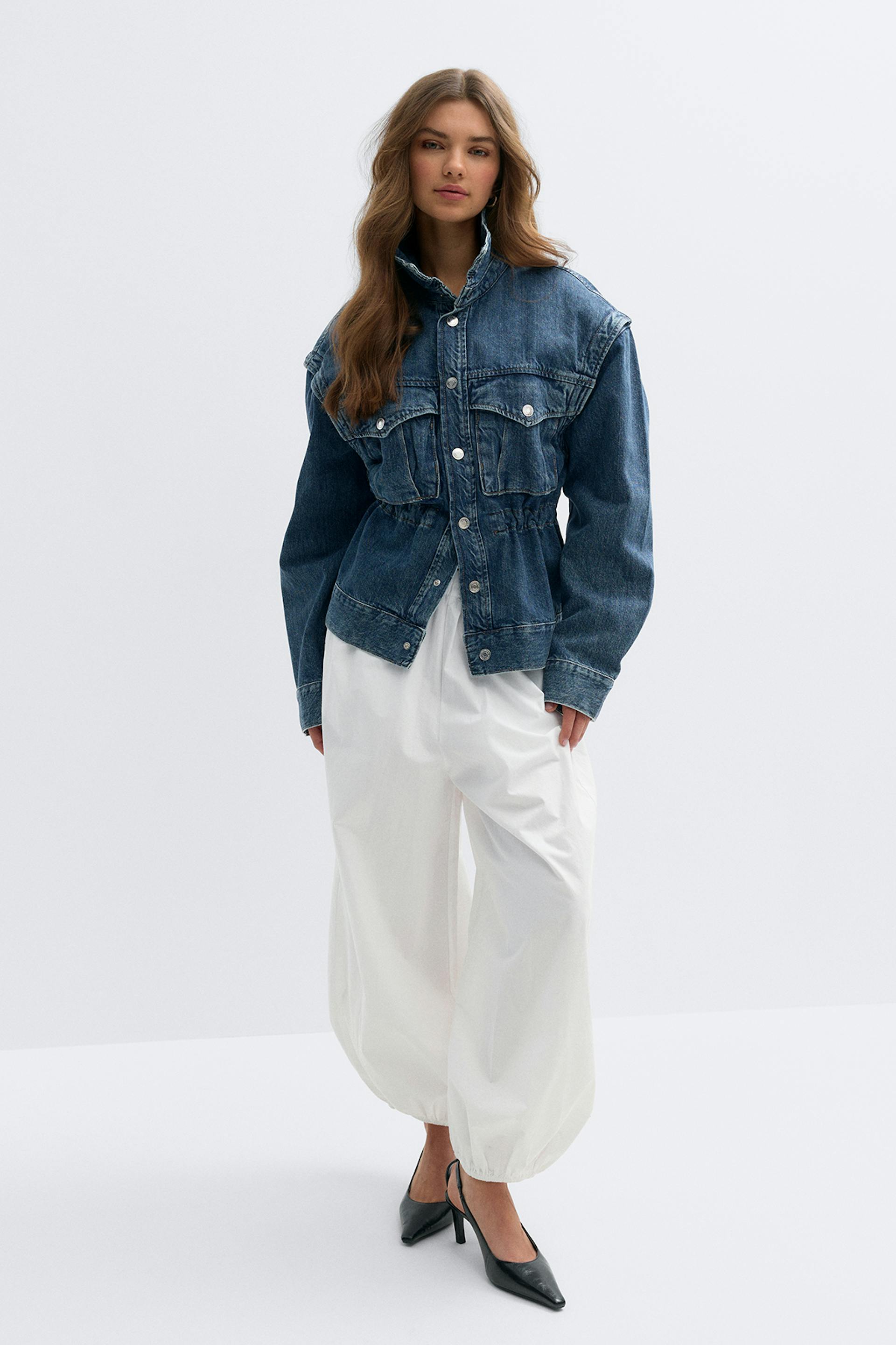 Dark blue denim jacket with high collar, snap-button front, flap chest pockets and cinched waist; white wide-leg balloon trousers with elasticized hems; black pointed-toe slingback heels.