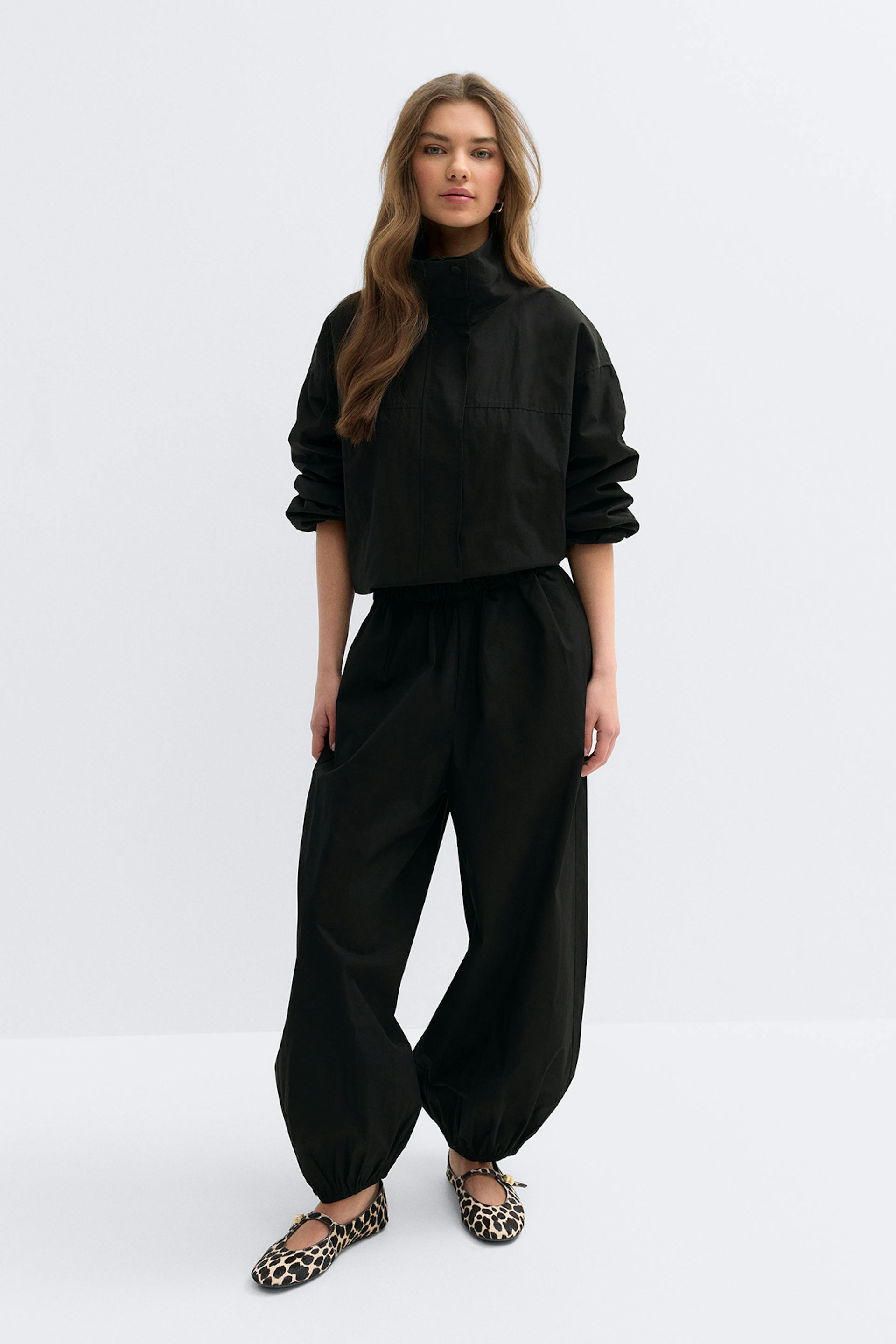 Black high-neck cropped jacket with long sleeves, matching black wide-leg balloon trousers with elasticated ankles, and leopard-print Mary Jane flats with bow detailing.