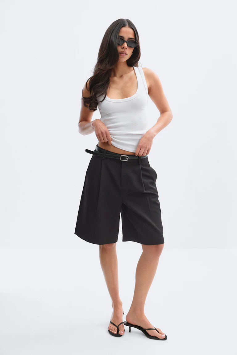 Weißes Tank-Top mit Rundhalsausschnitt, gepaart mit schwarzen, knielangen, maßgeschneiderten Bermudashorts, die mit einem dünnen schwarzen Gürtel zusammengehalten werden, einer schmalen schwarzen Sonnenbrille und schwarzen Zehensandalen mit Absatz.