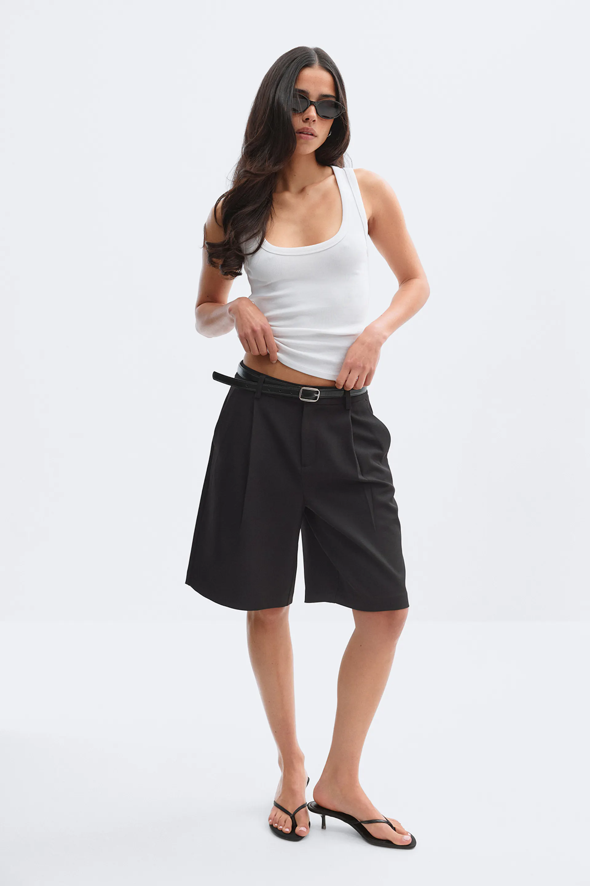 Hvid tanktop med rund hals kombineret med sorte knælange skræddersyede bermudashorts med et tyndt sort bælte, smalle sorte solbriller og sorte tangasandaler med hæl.