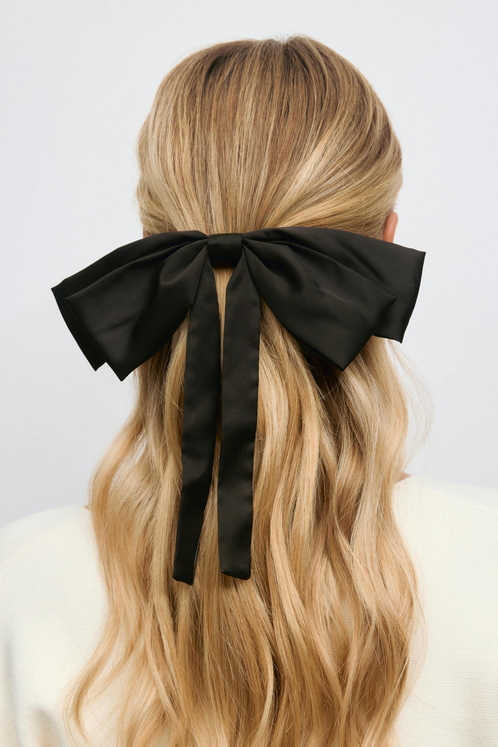 Black satin oversized bow hair accessory securing a half-up style, paired with a cream-colored knit top.