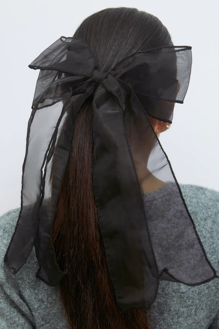Haarstrik van zwarte organza met lange, slepende uiteinden, vastgebonden over een lage paardenstaart; grijze gebreide trui met structuur.